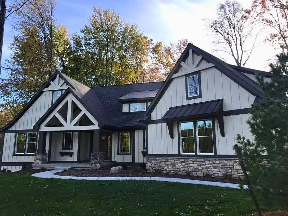 Front view of a Michigan, modern house with white vertical siding, black trim, stone accents, and a dark roof, set against a backdrop of trees and a partly cloudy sky.