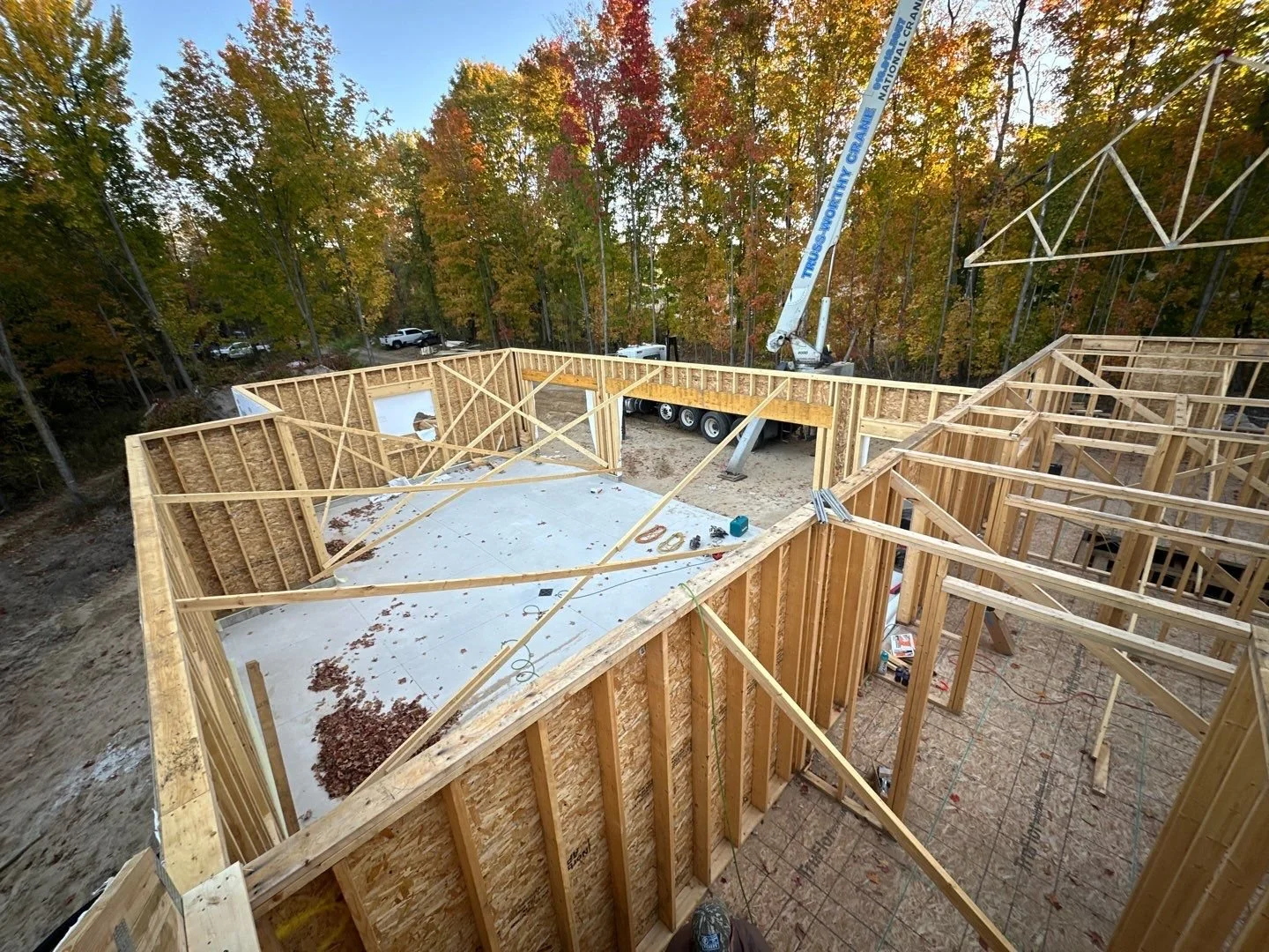 Construction site with wooden framing for a building, construction tools on the floor and a crane lifting materials. Autumn trees with colorful leaves are visible in the background.