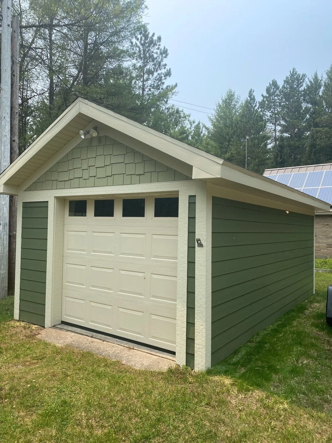 A detached garage with green horizontal siding and a white sectional garage door with small windows at the top, situated on a grassy yard with trees and power lines in the background. Custom James Hardie Color, Hardie Siding, trim, & Shakes. Quality