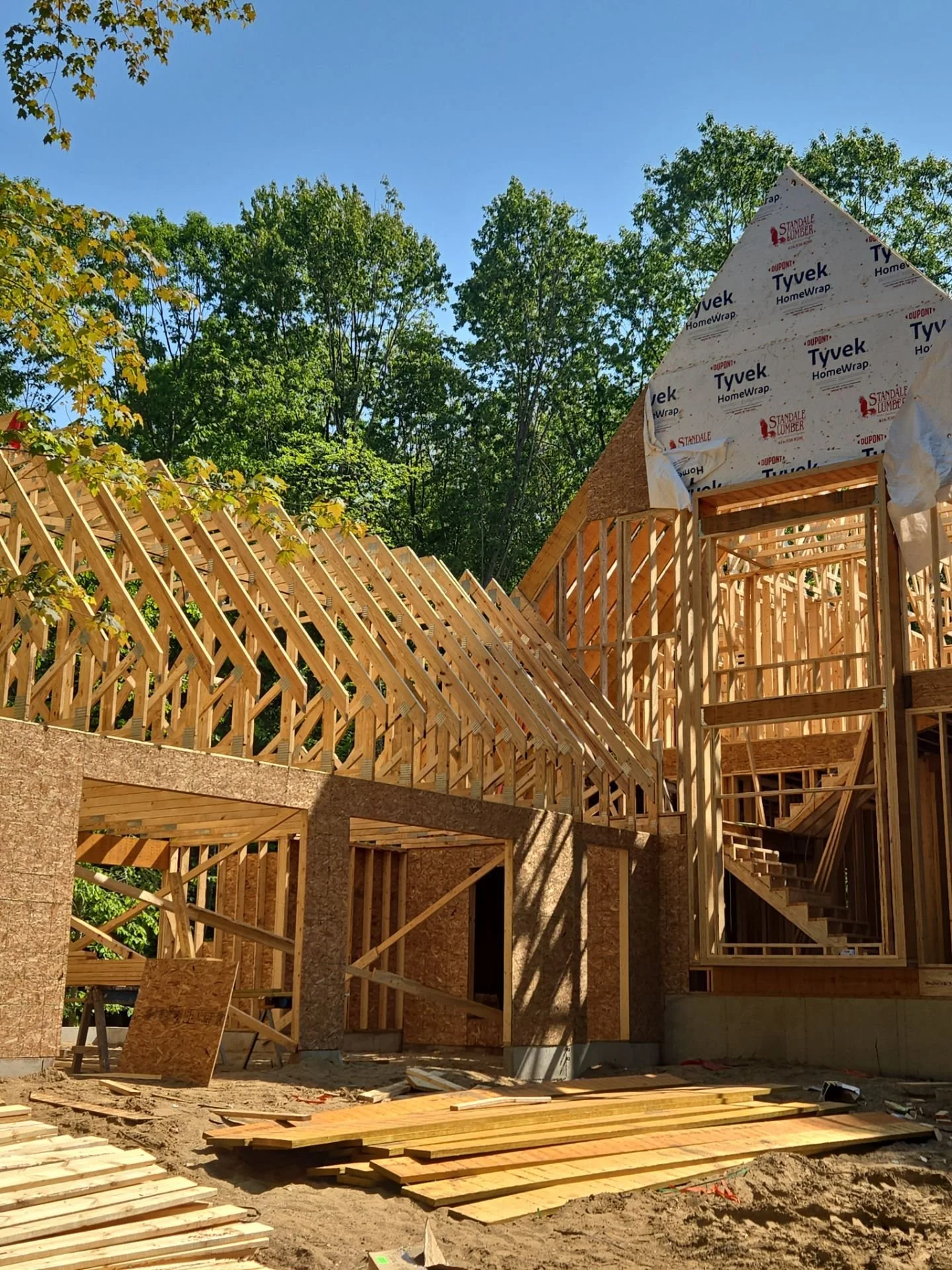 Wooden framing of a house under construction with blue sky and green trees in the background.