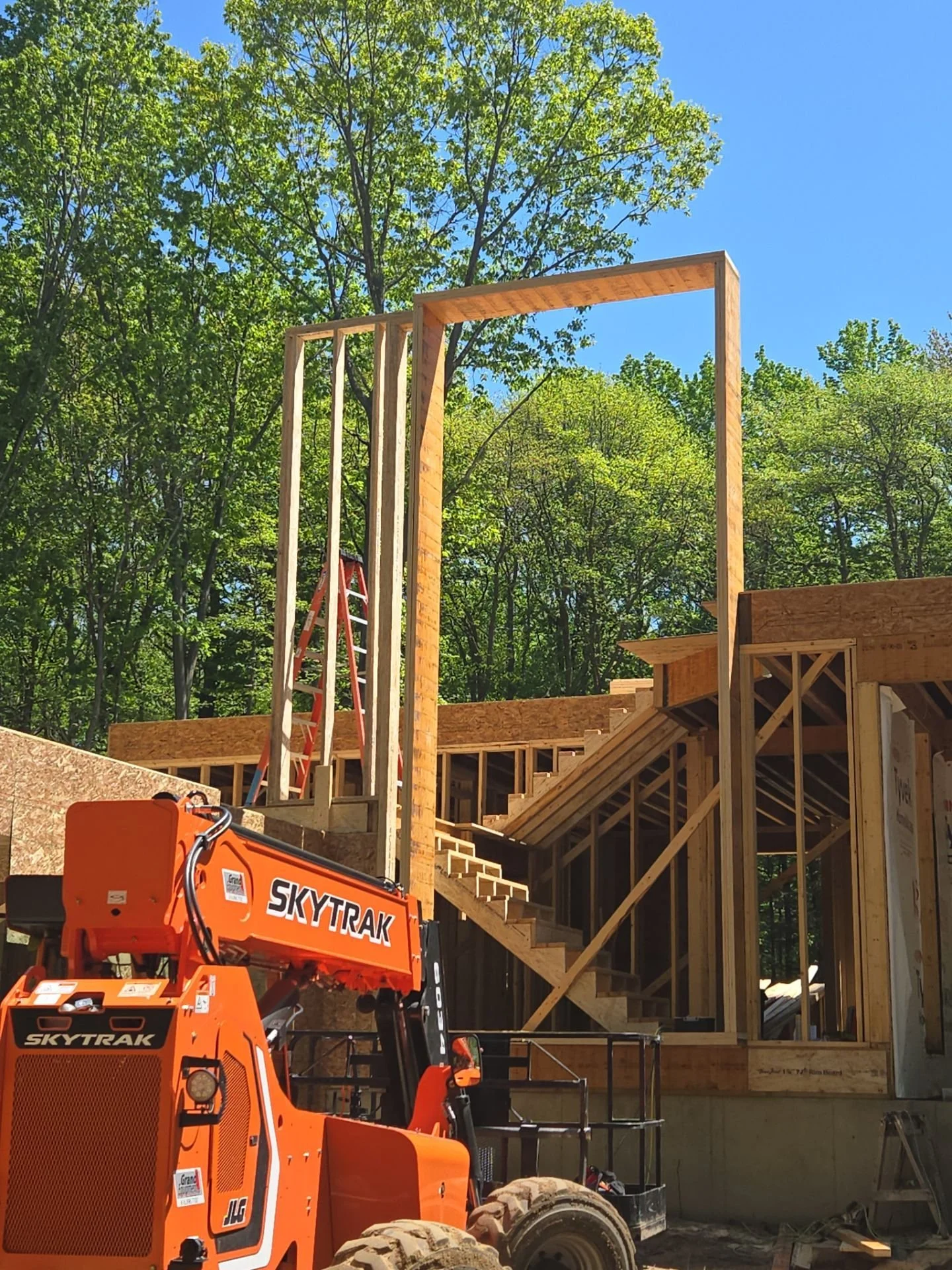 Construction site with wooden framing and stairs, orange SkyTrak lift, green trees, and blue sky.