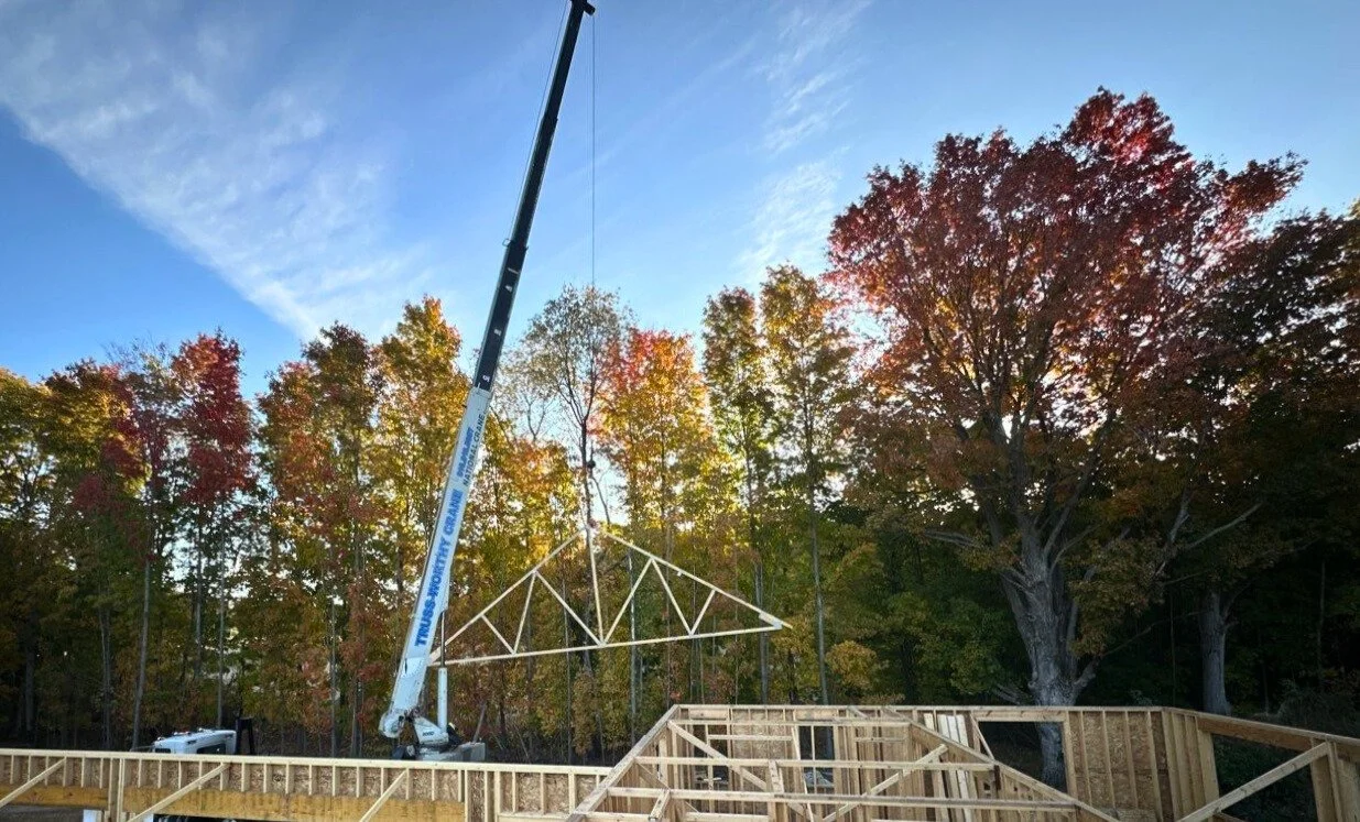A construction site with a crane lifting a wooden roof truss into place, surrounded by trees with autumn leaves.