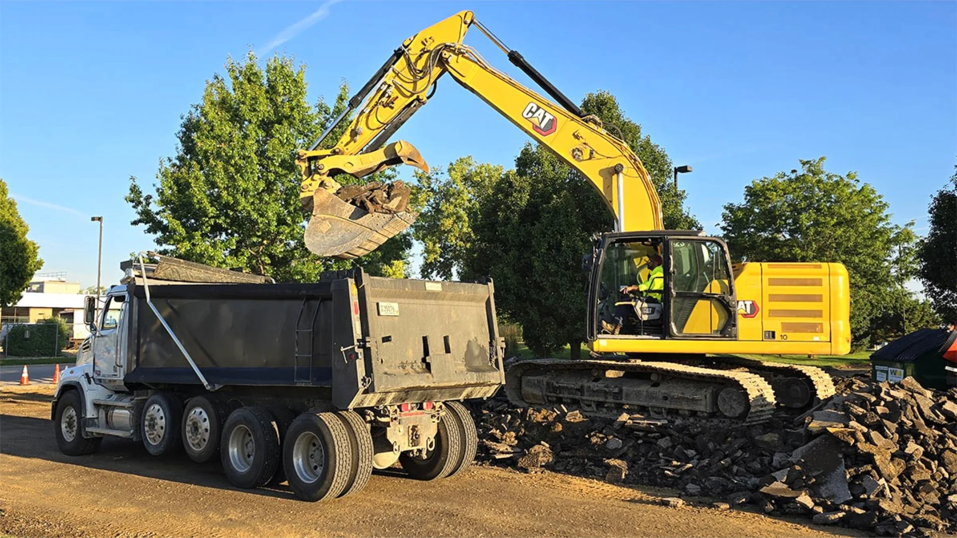 Excavator loading debris into a dump truck at a construction site, surrounded by trees and clear blue sky.
