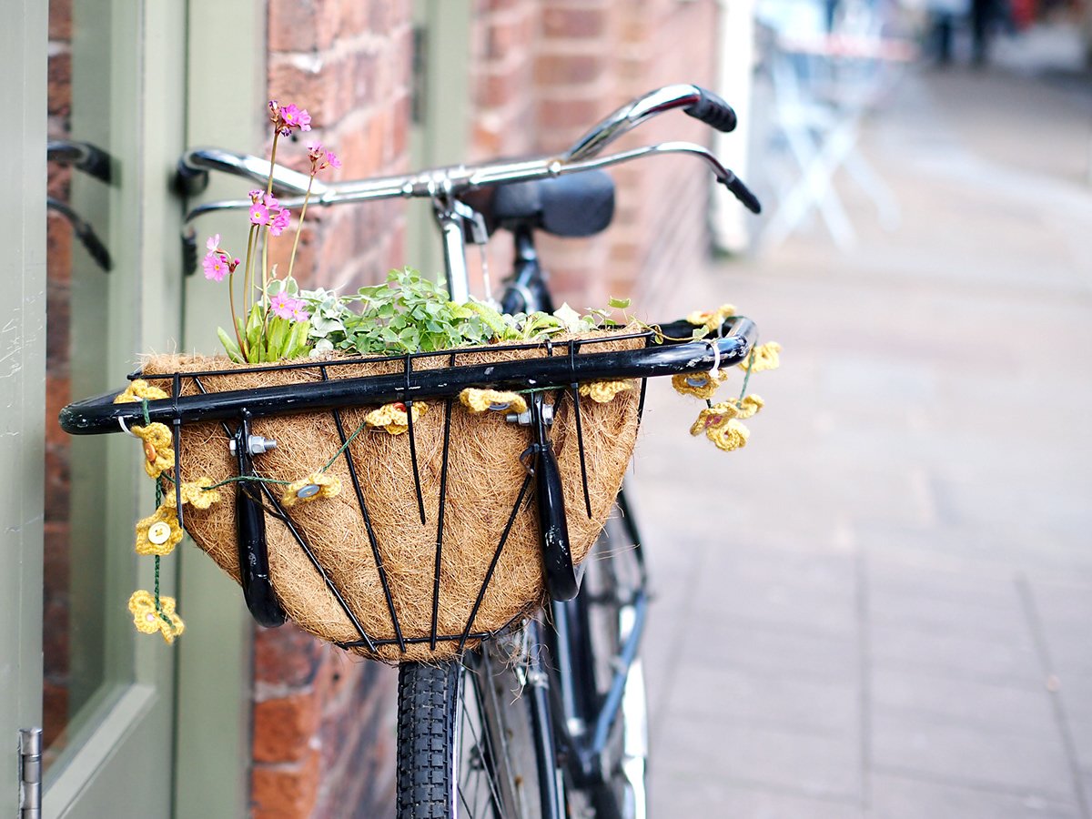 A bicycle with a front basket filled with colorful flowers and greenery, parked on a sidewalk next to a brick building.