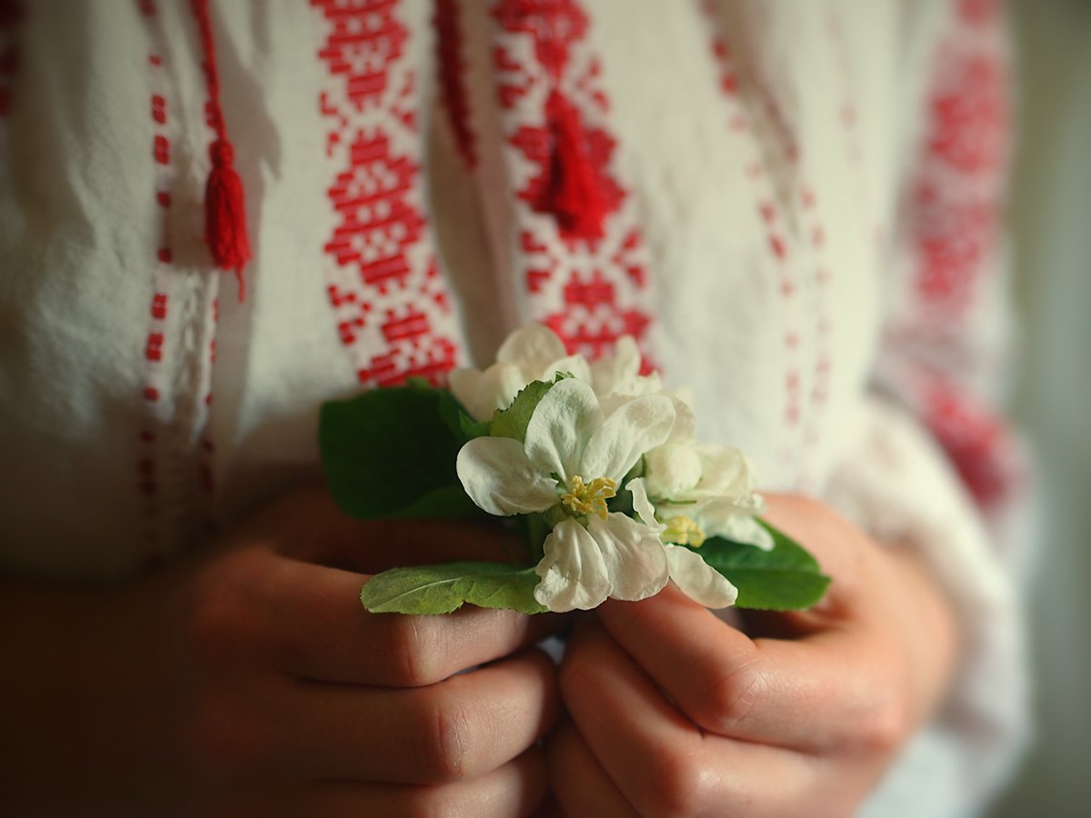 Person holding white flowers and green leaves, wearing a traditional embroidered garment with red patterns.