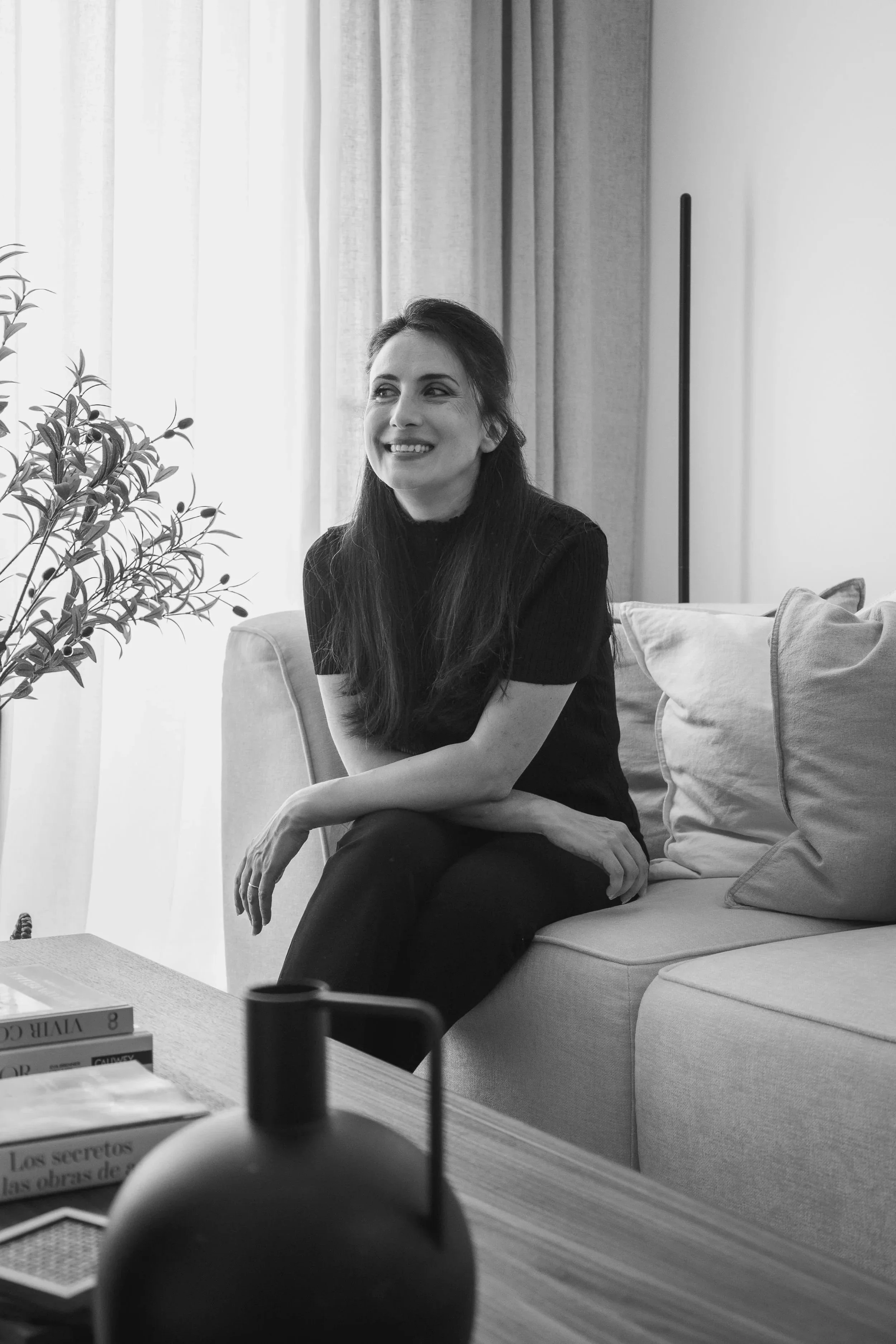 Mujer sonriente sentada en un sillón en una sala con cortinas y plantas, vista en escala de grises.