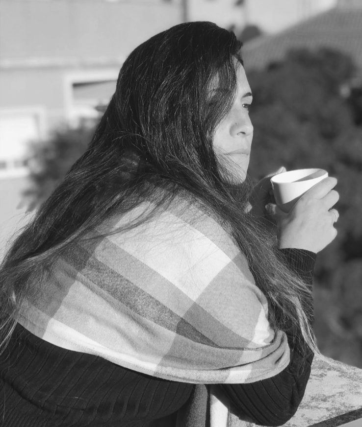 Mujer con cabello largo y oscuro, sosteniendo una taza, con una bufanda a cuadros y una expresión pensativa.