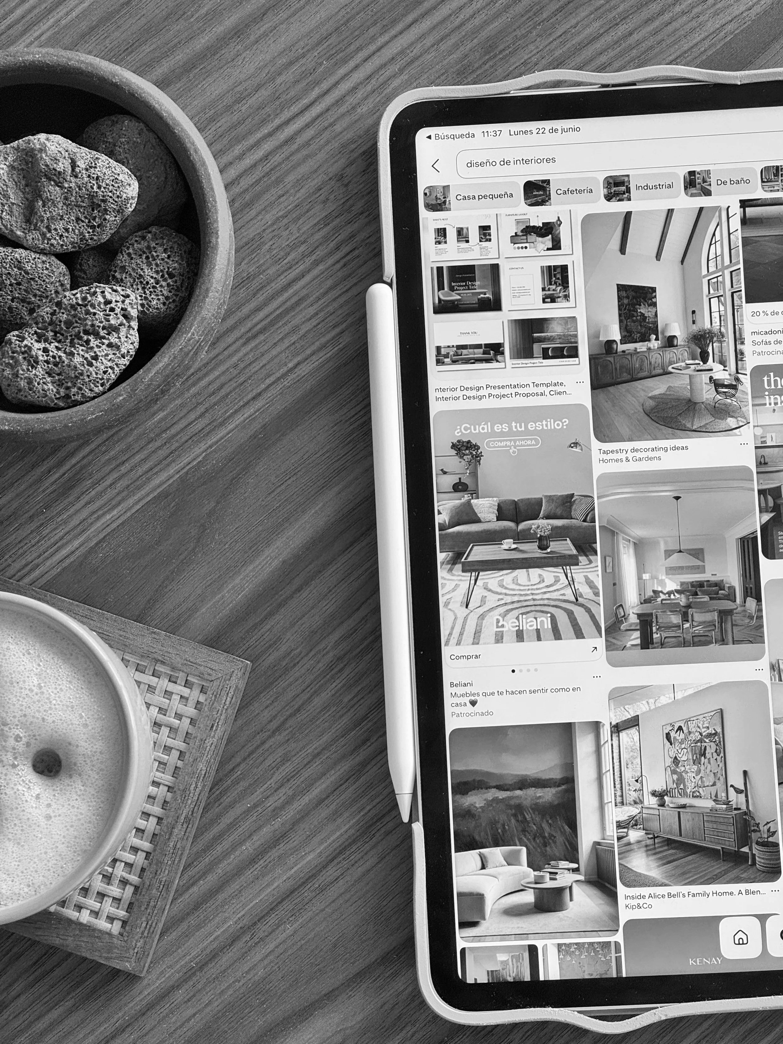 La imagen muestra una tableta en una superficie de madera con un bol de piedras volcánicas y una pequeña bandeja de ratán con una taza de café. La pantalla de la tableta tiene una búsqueda en línea sobre diseños de interiores y muestra varias imágenes de salas y proyectos de decoración.