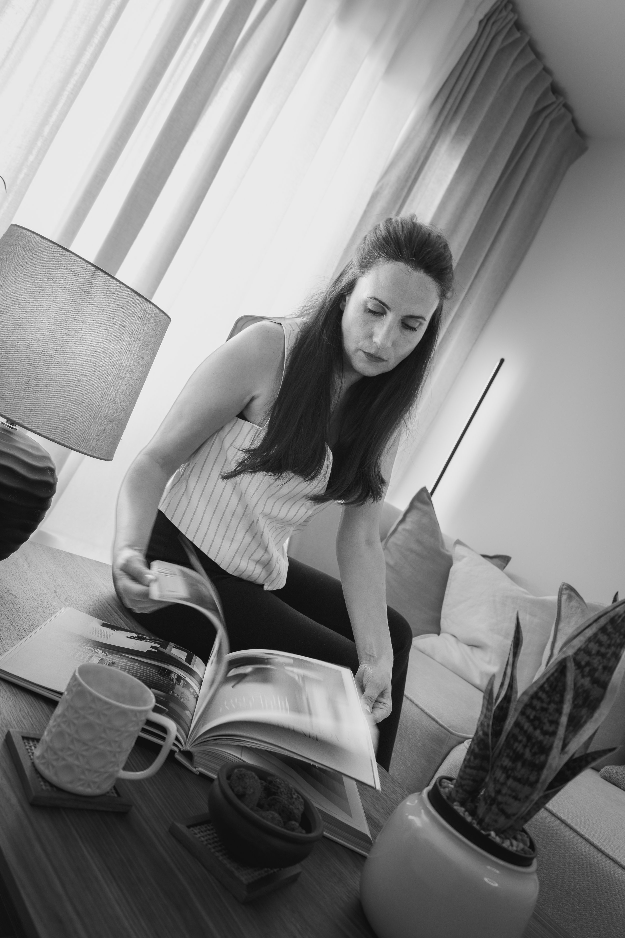 Mujer con cabello largo sentado en una mesa con libros, tazas y recipientes con comida, en un ambiente interior con cortinas blancas.