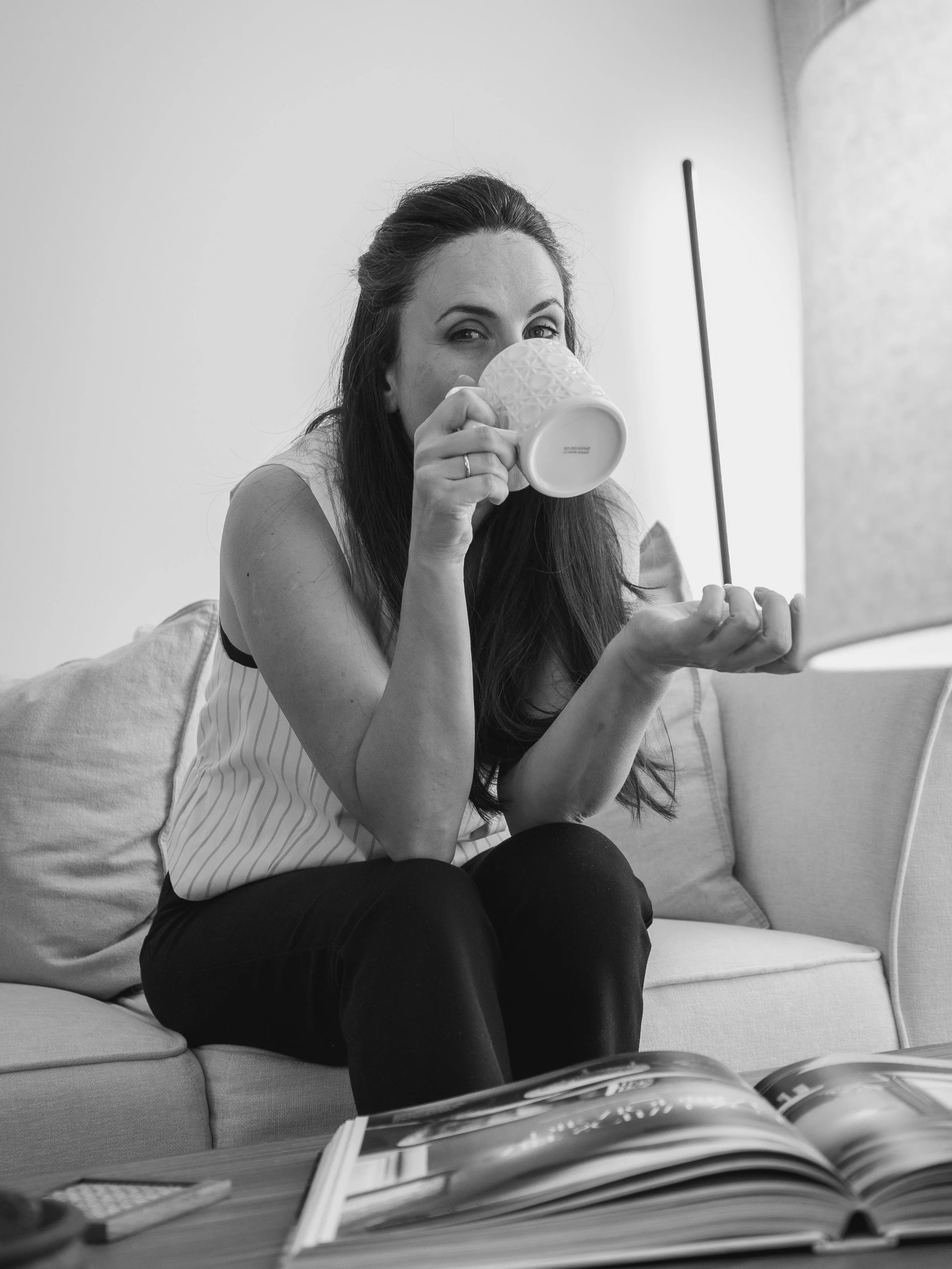 Una mujer con cabello largo y oscuro, que está sentada en un sofá, tomando una taza y sosteniendo un tubo con un dedo, en un entorno interior en blanco y negro.