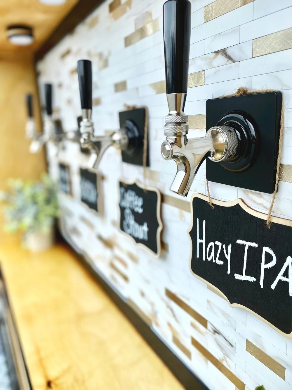 A row of beer taps with black handles and chalkboard labels. The labels read different beer types, such as "Hazy IPA." The background features a tiled wall with a light wood bar top.