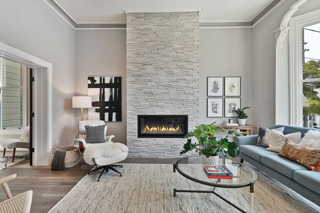 Modern living room with a stone fireplace, a gray sofa with cushions, a glass coffee table, a white armchair with a sheepskin throw, a side table with a plant, and a window with a view of trees.