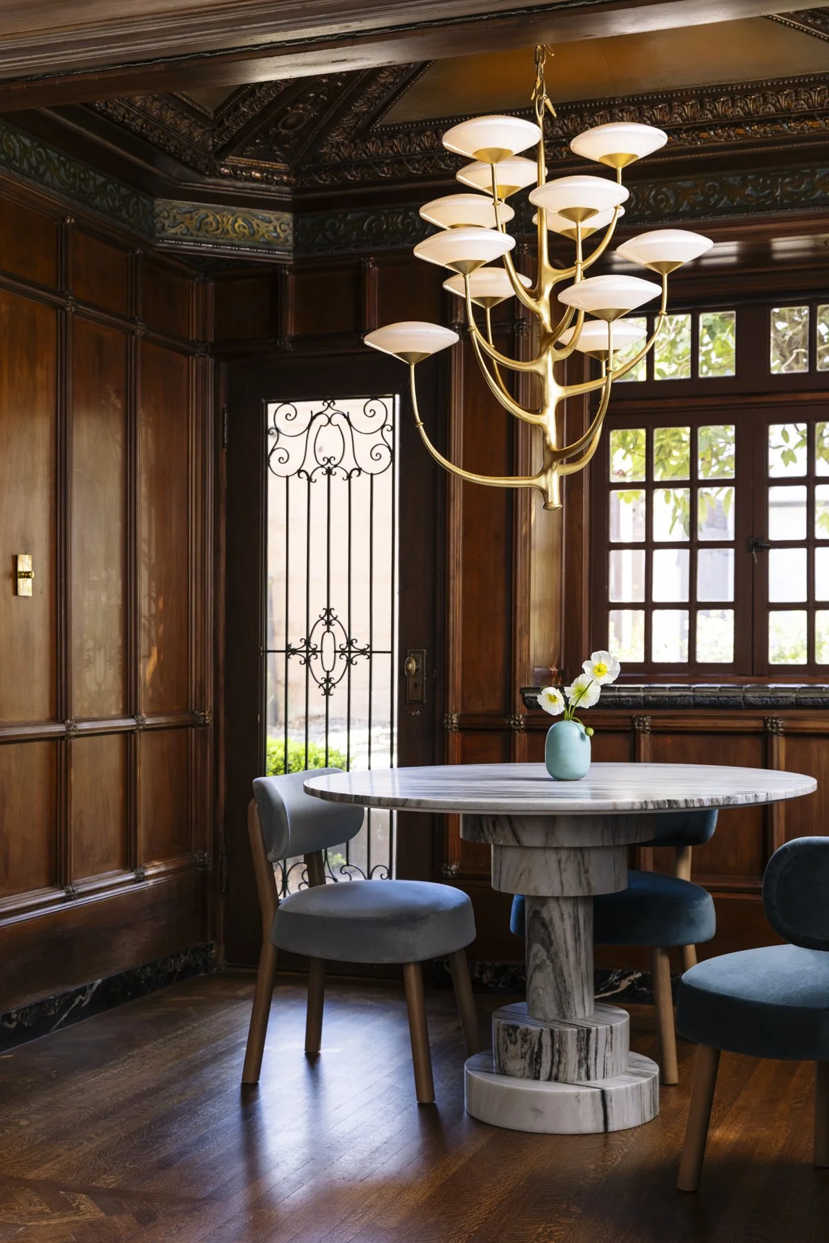 Luxury dining room renovation in a historic San Francisco home featuring original wood paneling, a sculptural brass chandelier, a round marble dining table, velvet dining chairs, and large windows that fill the space with natural light.