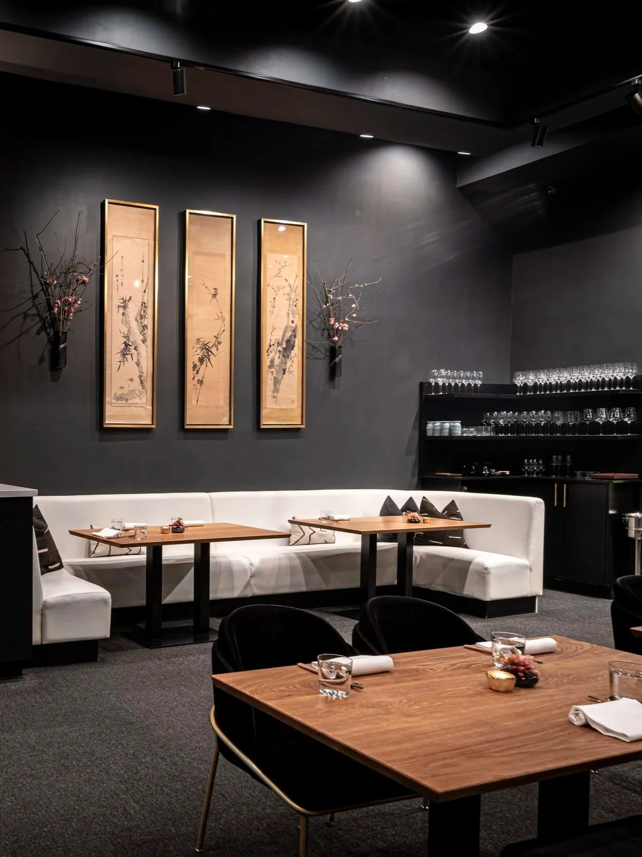 Interior of an elegant restaurant with dark walls, Japanese scrolls and floral decorations, white seating, and wooden tables set for dining.