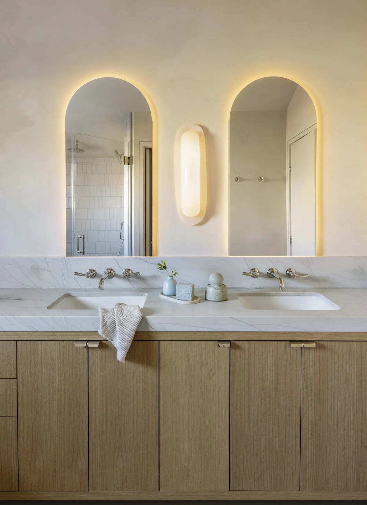 Luxury bathroom renovation with a double vanity, white marble countertop, white oak cabinetry, arched backlit mirrors, integrated wall lighting, and contemporary fixtures in a custom San Francisco home.