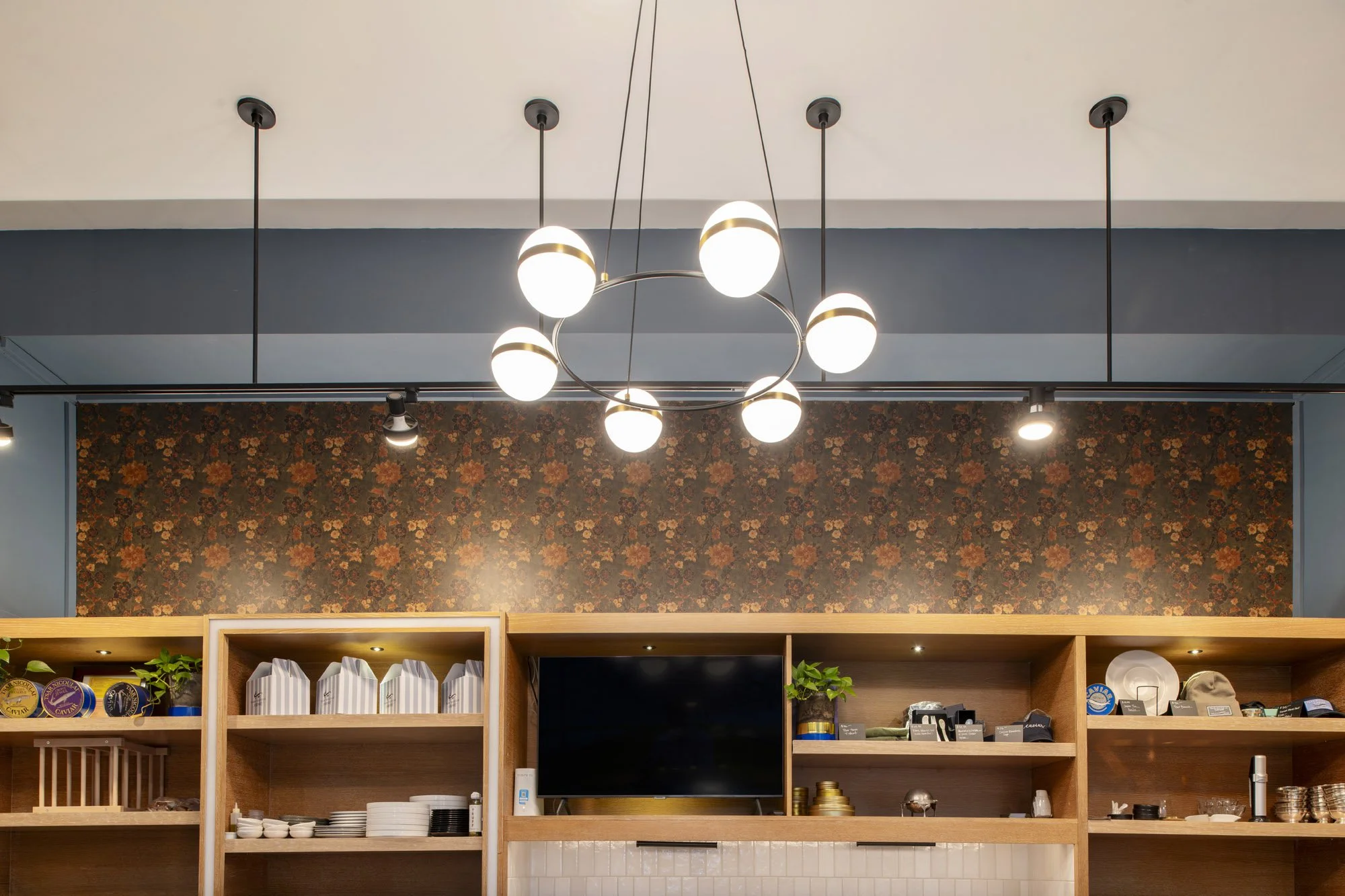 Indoor kitchen shelving with a mounted TV, shelves with plates, cups, and decorative items, hanging modern chandelier with globe lights, background with floral patterned wallpaper with dark blue walls.