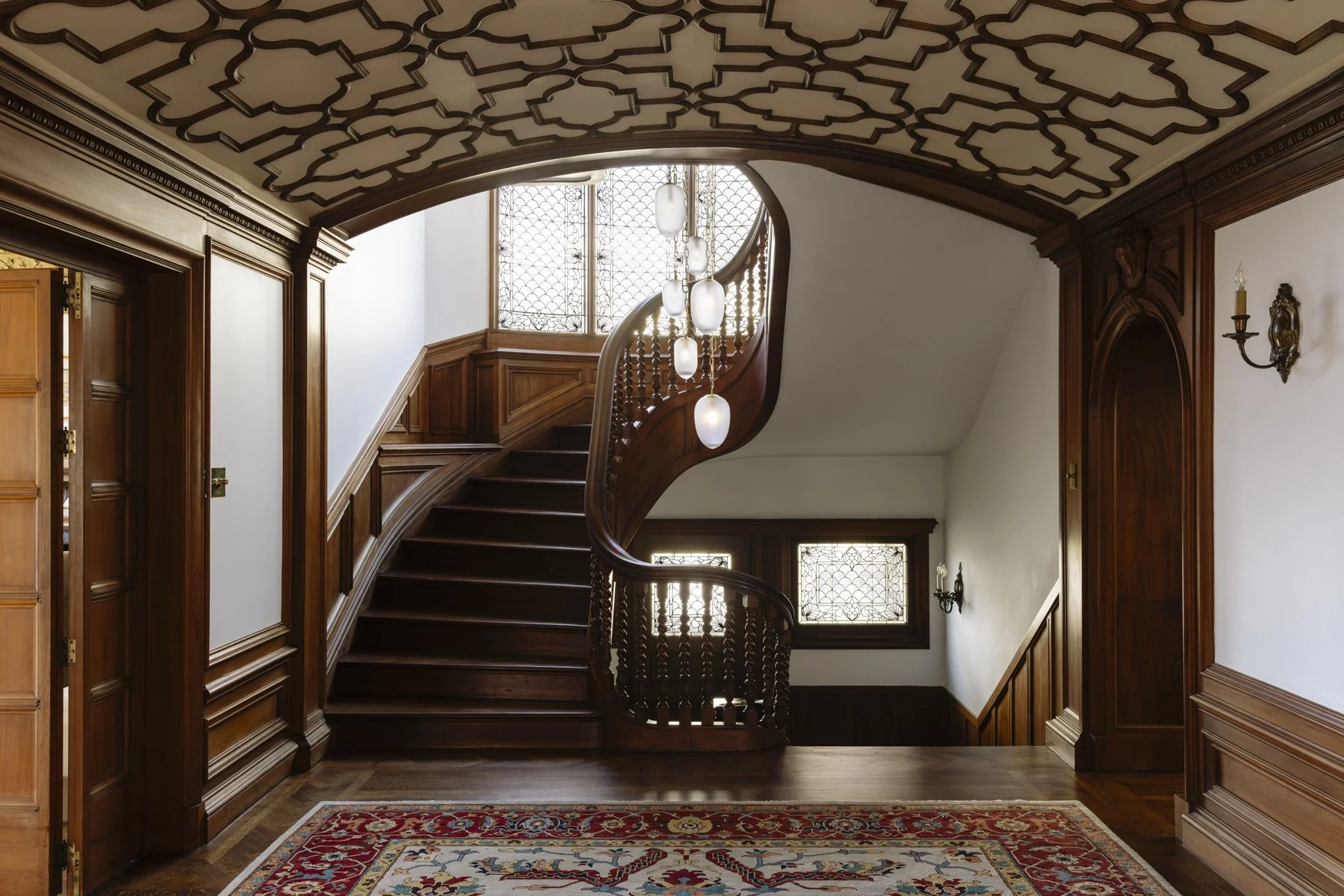 Luxury residential staircase featuring custom wood railings, intricate ceiling millwork, and natural light from stained glass windows.