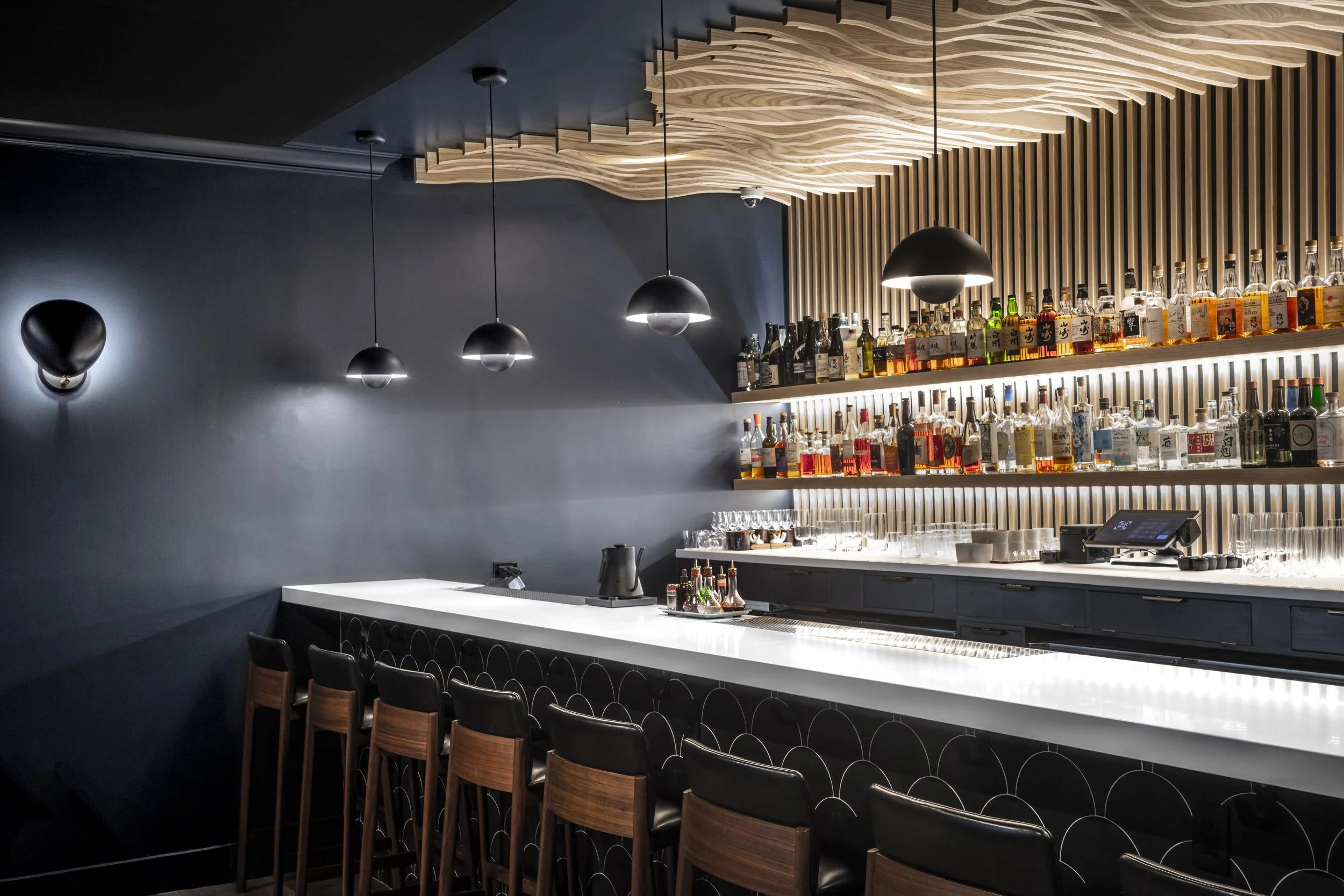 Bar interior with a white counter, wooden chairs, a display of liquor bottles on shelves, and modern black pendant lights hanging from a wooden wave-designed ceiling.