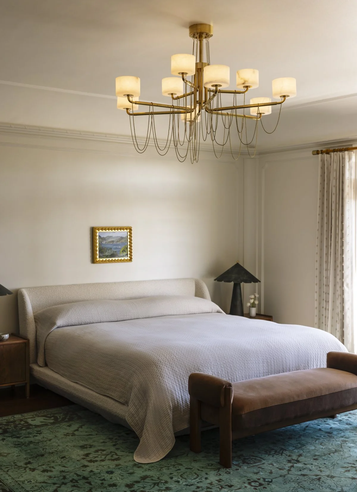 Luxury primary bedroom featuring a curved upholstered bed, custom brass chandelier, warm neutral palette, designer furnishings, and large windows with natural light in a renovated San Francisco home.