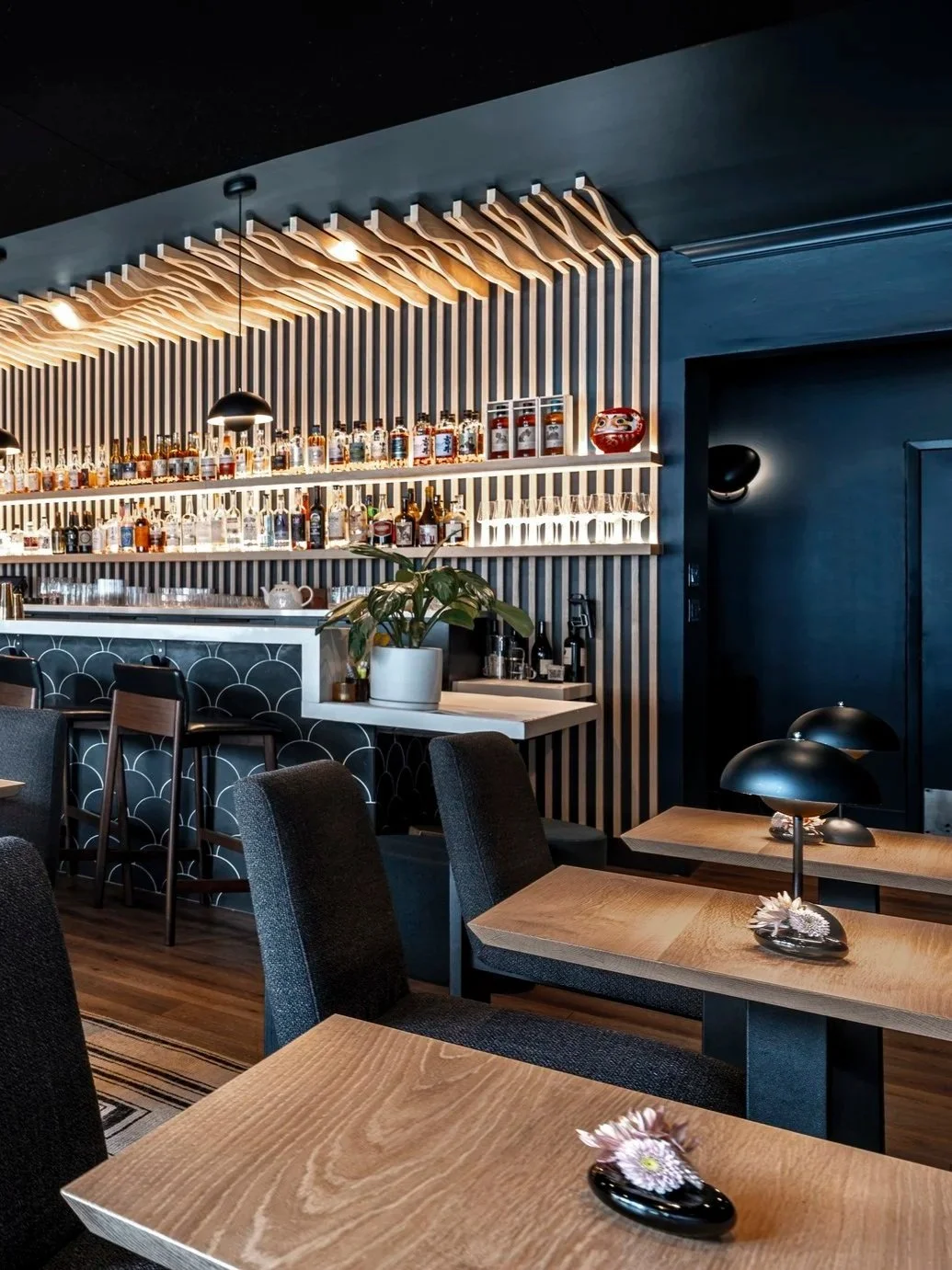 Modern restaurant interior with wooden tables, dark chairs, a bar area with various bottles, and decorative lighting in a stylish, dimly-lit setting.