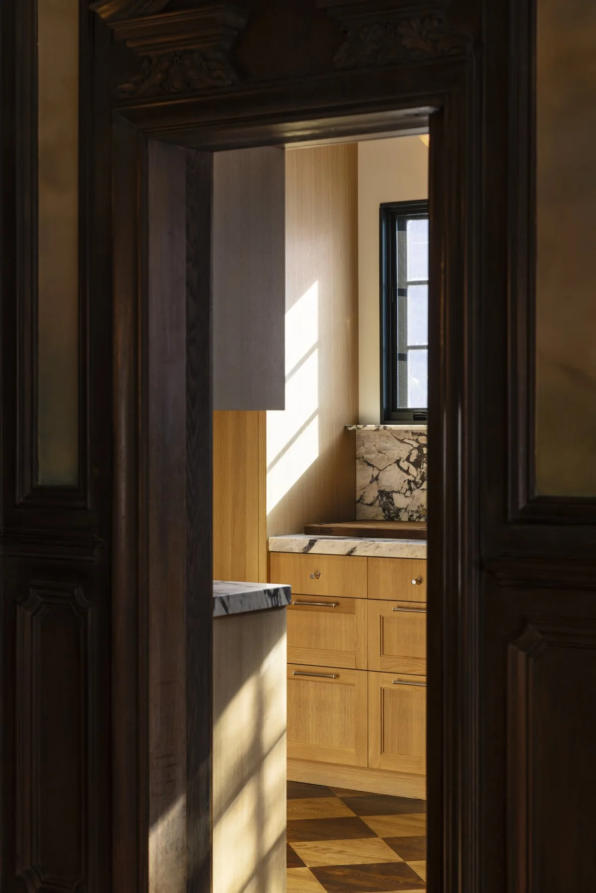 View through a restored wood doorway into a luxury custom kitchen in a historic San Francisco home, featuring white marble countertops, natural oak cabinetry, a black-framed window, and warm natural light.