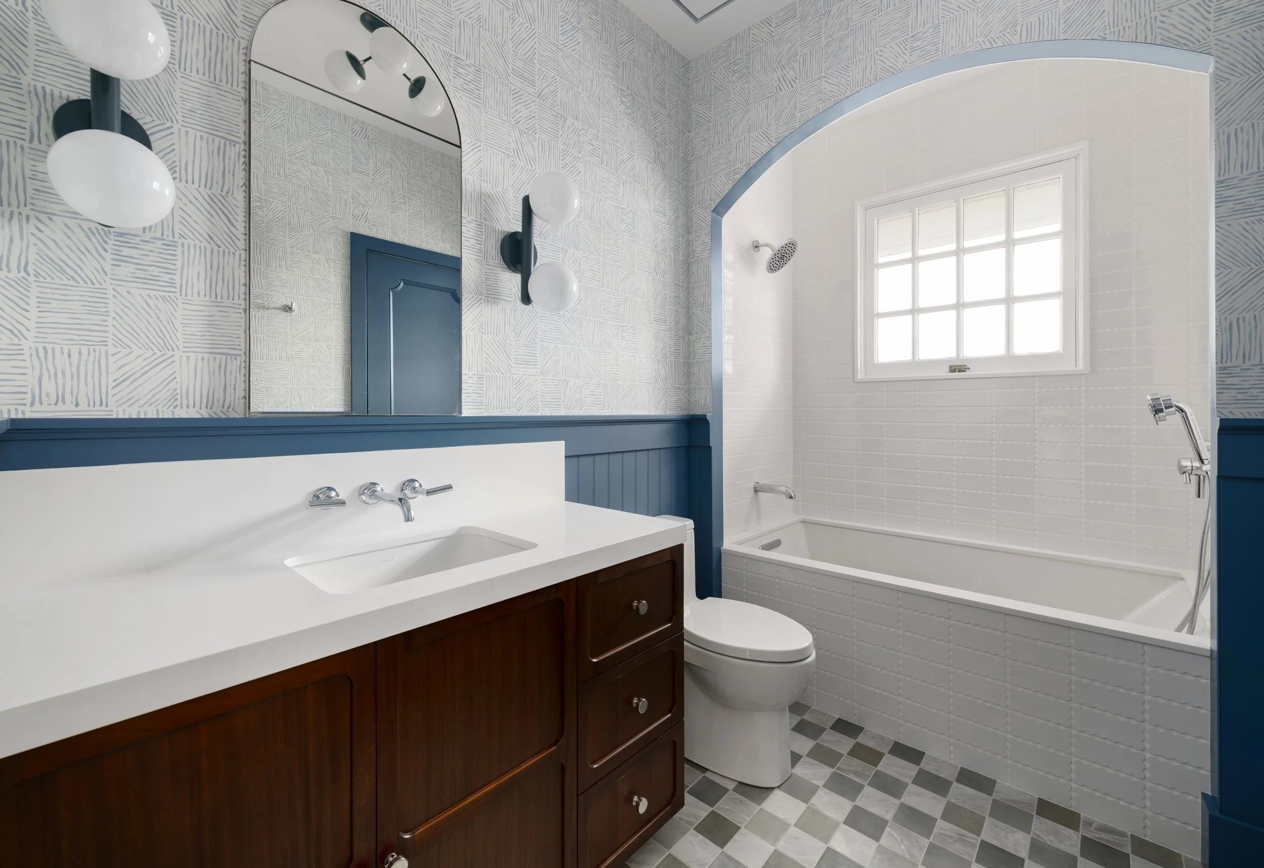 A modern bathroom with white tiled walls and a large window. There is a white bathtub, a toilet, a wooden vanity with a white countertop, and a large mirror with light fixtures above it. The ceiling and trim are painted in light blue.