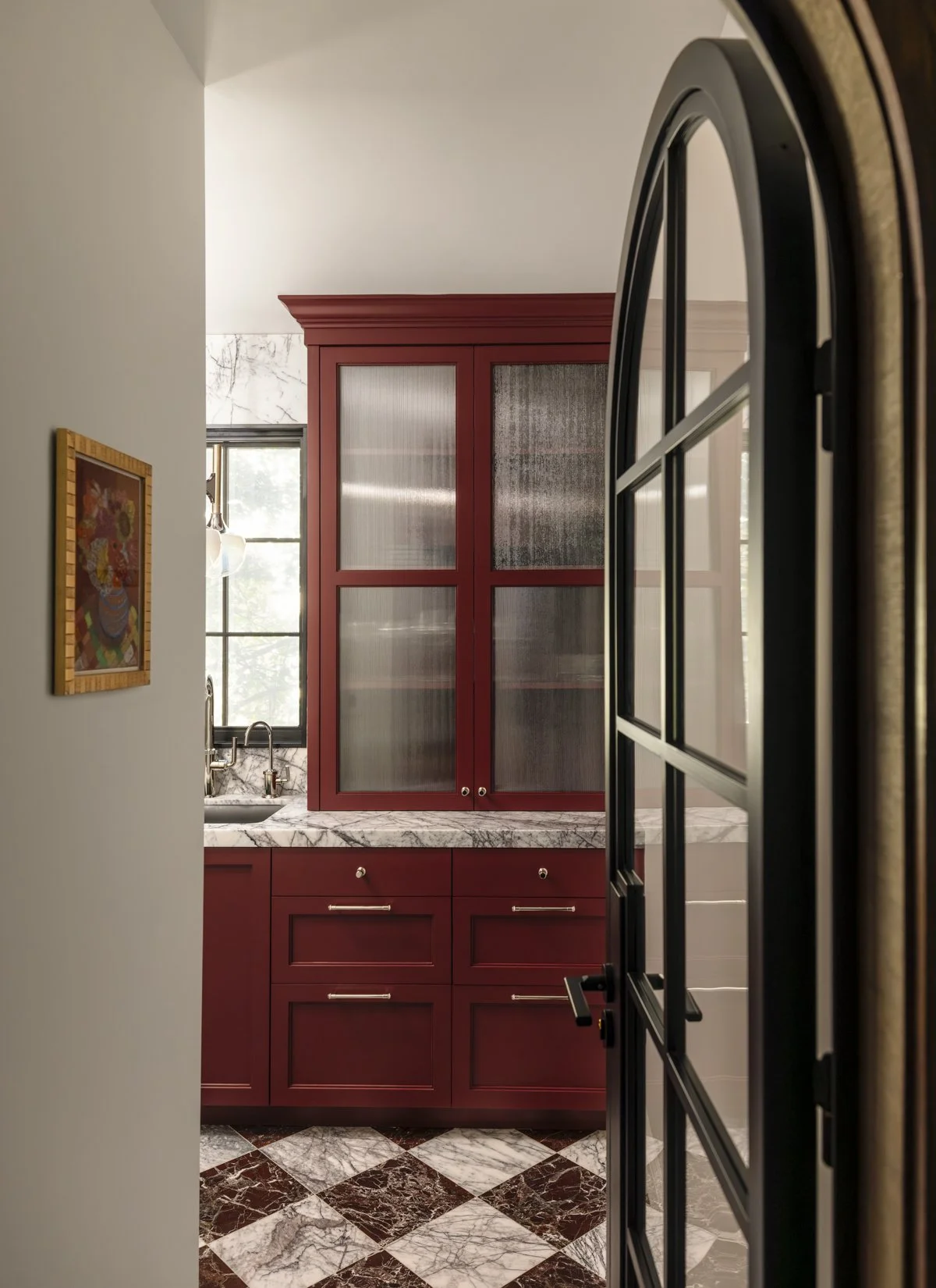 Custom butler’s pantry in a luxury San Francisco home featuring deep red custom cabinetry, marble countertops, textured glass cabinet doors, a black steel-framed glass door, and natural light from a nearby window.