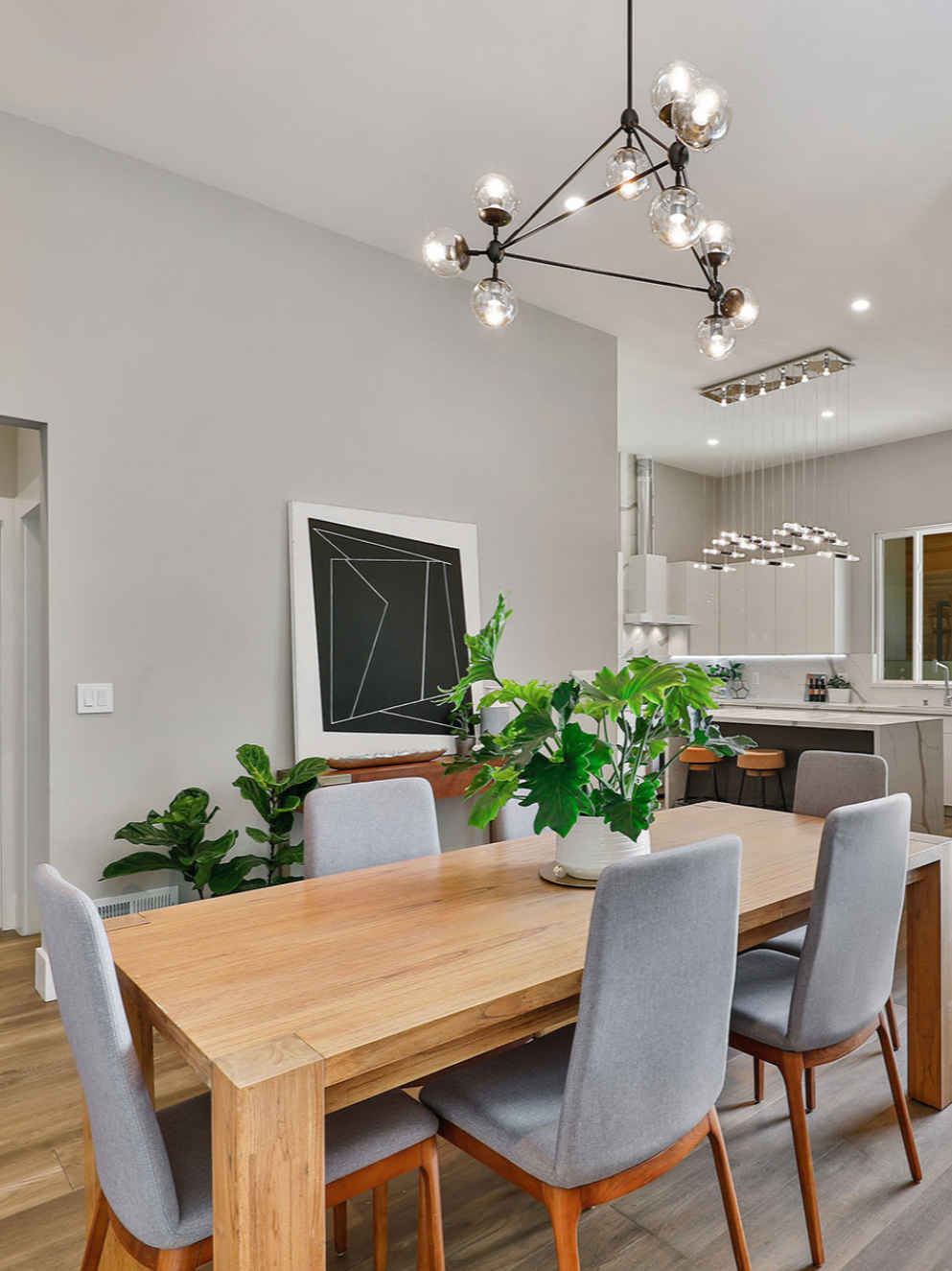 Modern dining room with a wooden table, gray upholstered chairs, a large potted plant, abstract black and white artwork on the wall, and contemporary light fixtures.