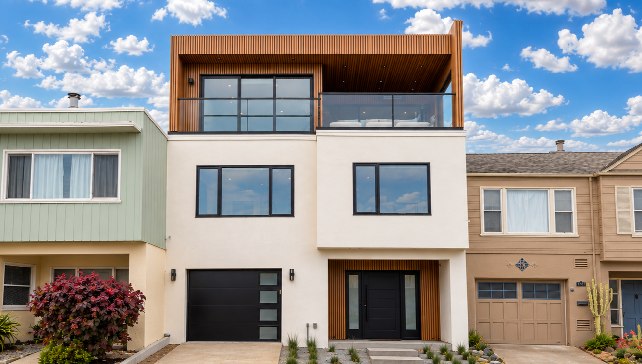 Modern multi-story house with a white facade, black window frames, and wooden accents near the entrance and on the top floor balcony. Front yard has plants and a paved walkway, neighboring houses on either side, and a partly cloudy sky in the background.