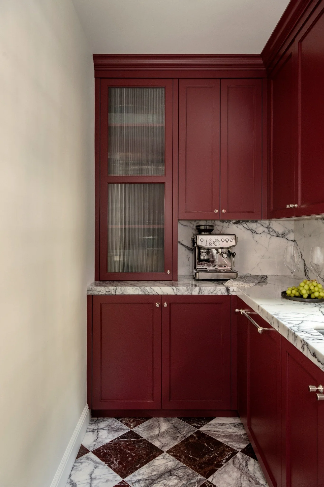 A kitchen corner with deep red cabinets, a marble countertop, and a stainless steel espresso machine. The floor has a classic black and white diamond pattern marble tile. There is a bowl of green grapes on the counter.