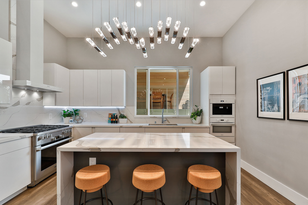 Modern kitchen with white cabinetry, marble countertops, a large kitchen island with three orange stools, a window above the sink, and a stylish chandelier overhead.