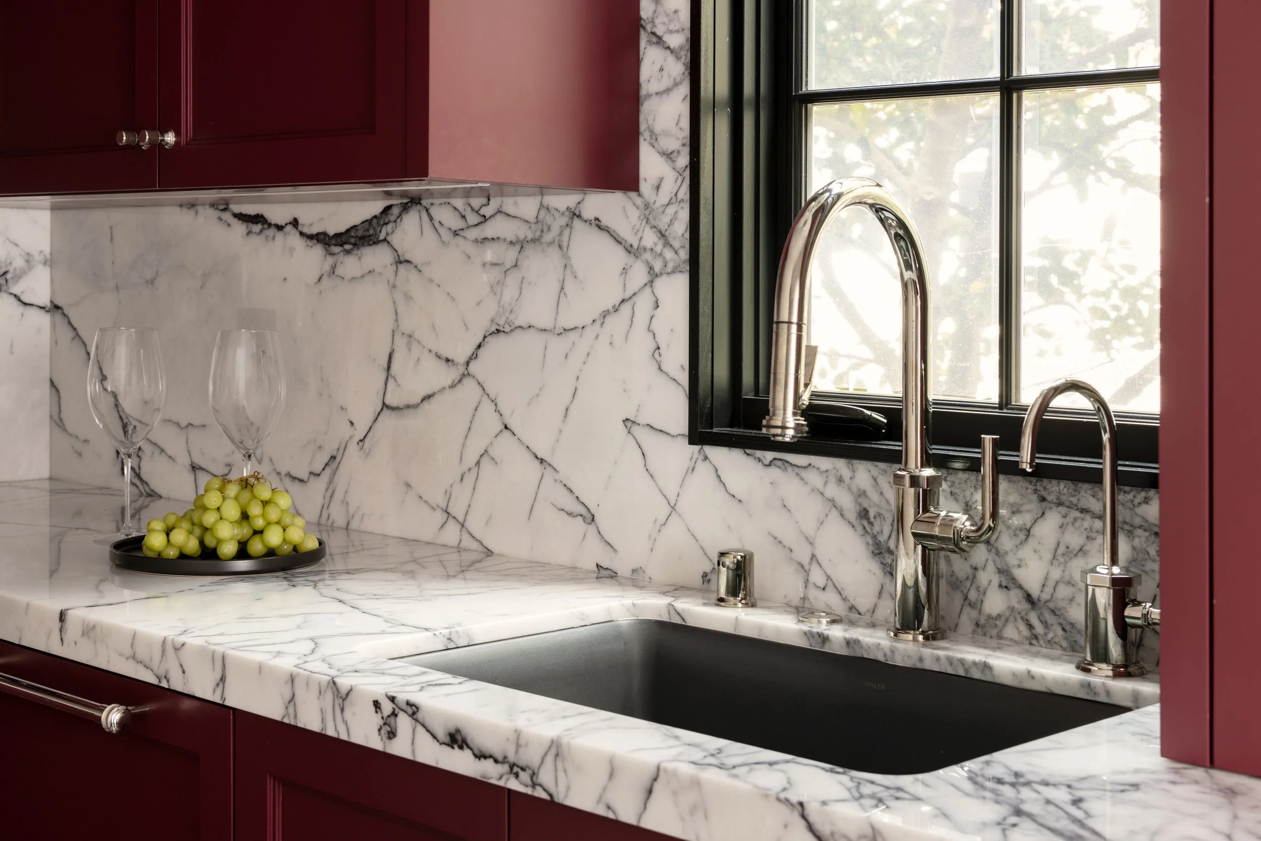 Kitchen counter with white marble surface, red cabinets, a black and white marble backsplash, a window with a black frame, a chrome kitchen faucet, two empty wine glasses, and a black tray of green grapes.