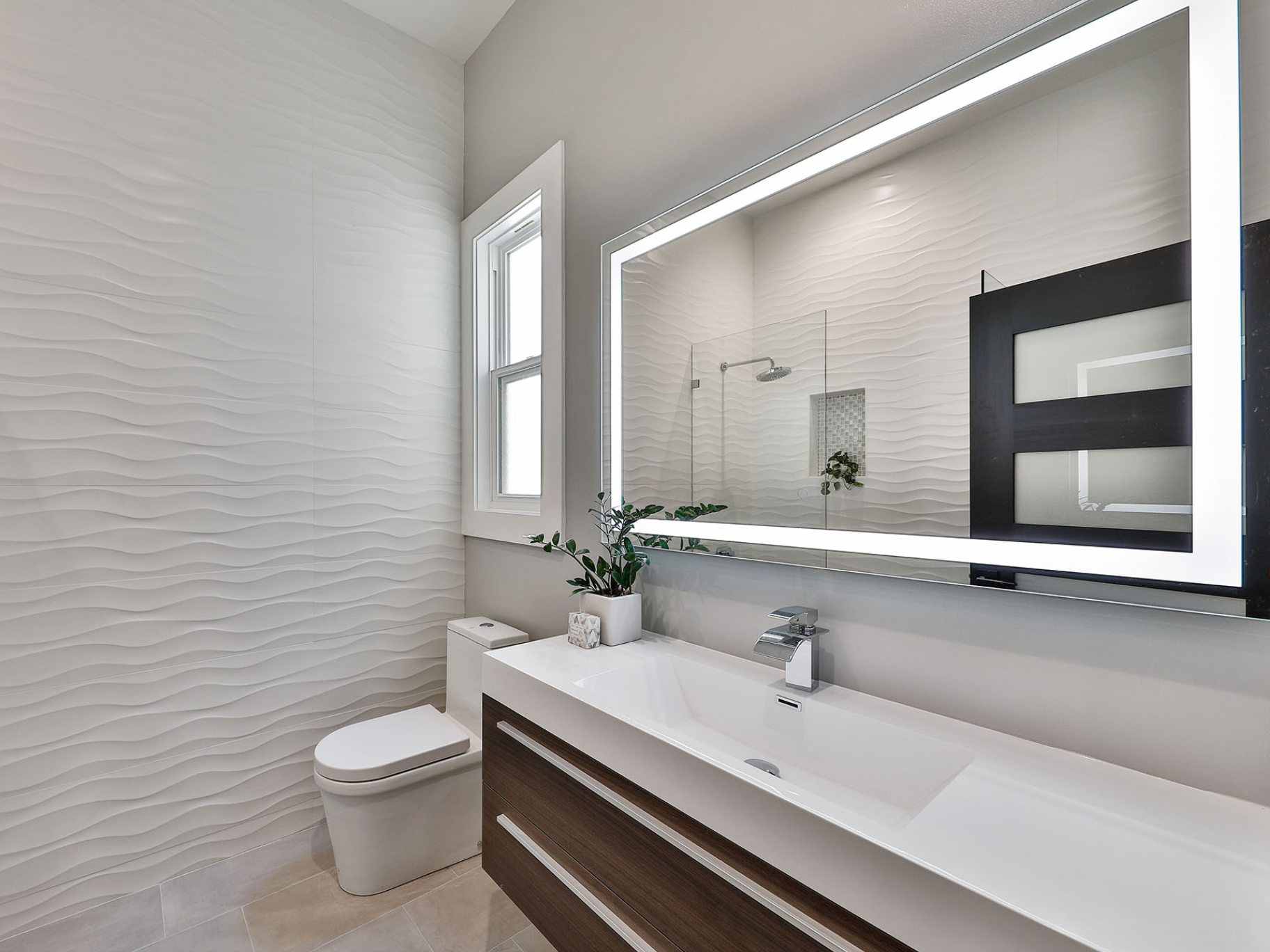 Modern bathroom with white textured wave-patterned wall tiles, a toilet, a white vanity with a dark wood drawer, a large illuminated mirror, a small potted plant, a window, and a shower with glass enclosure visible in the reflection.