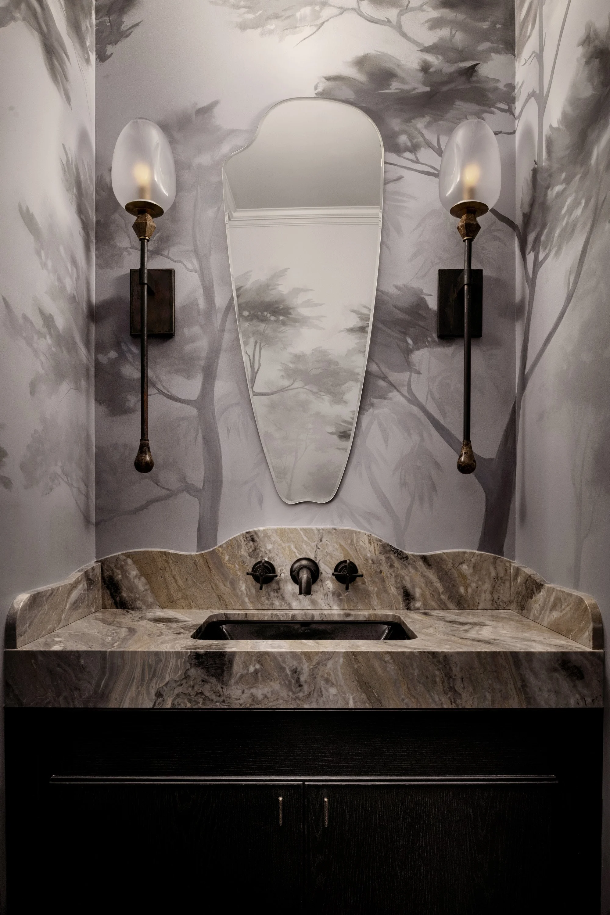 A bathroom vanity with a dark wooden cabinet, a marble countertop, and a built-in sink. Two wall-mounted sconces with frosted glass shades and bronze fixtures are on the wall. A decorative mirror with a curved top reflects a painted mural of trees in grayscale.