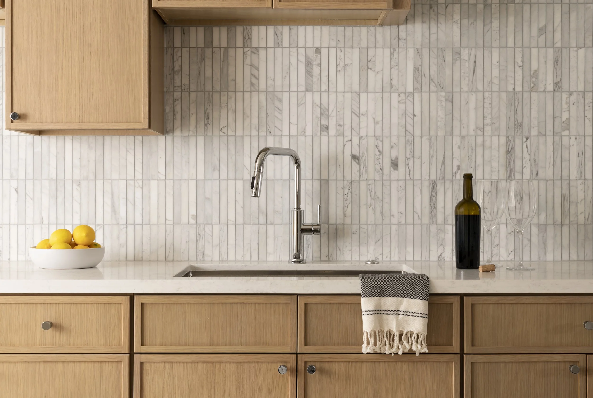 Kitchen with wooden cabinets, marble backsplash, a stainless steel faucet over a sink, a bottle of wine and two wine glasses on the counter, a bowl of lemons, and a white towel with black stripes and fringe hanging on the cabinet.