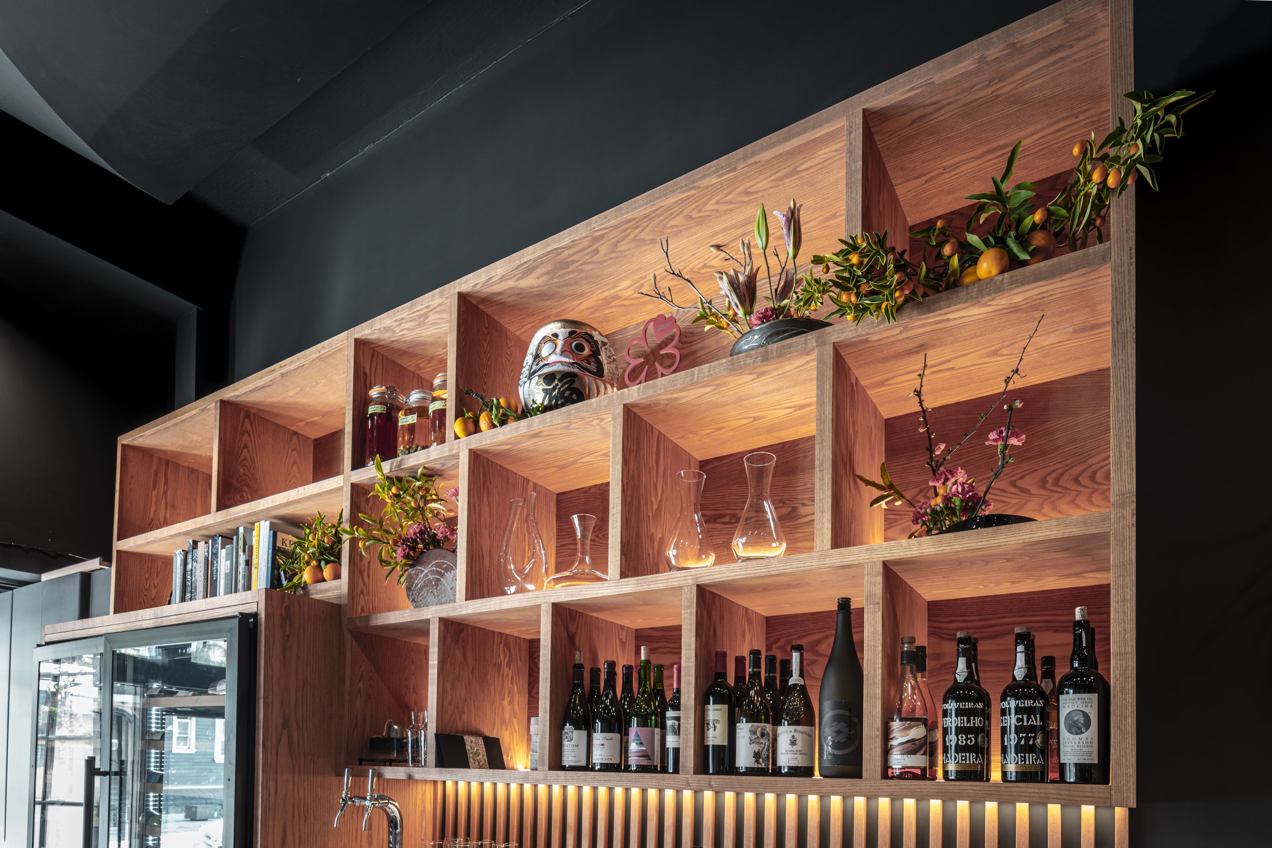A wooden bar shelving unit with decorative items, books, flowers, and bottles of wine and alcohol, illuminated by warm lighting.
