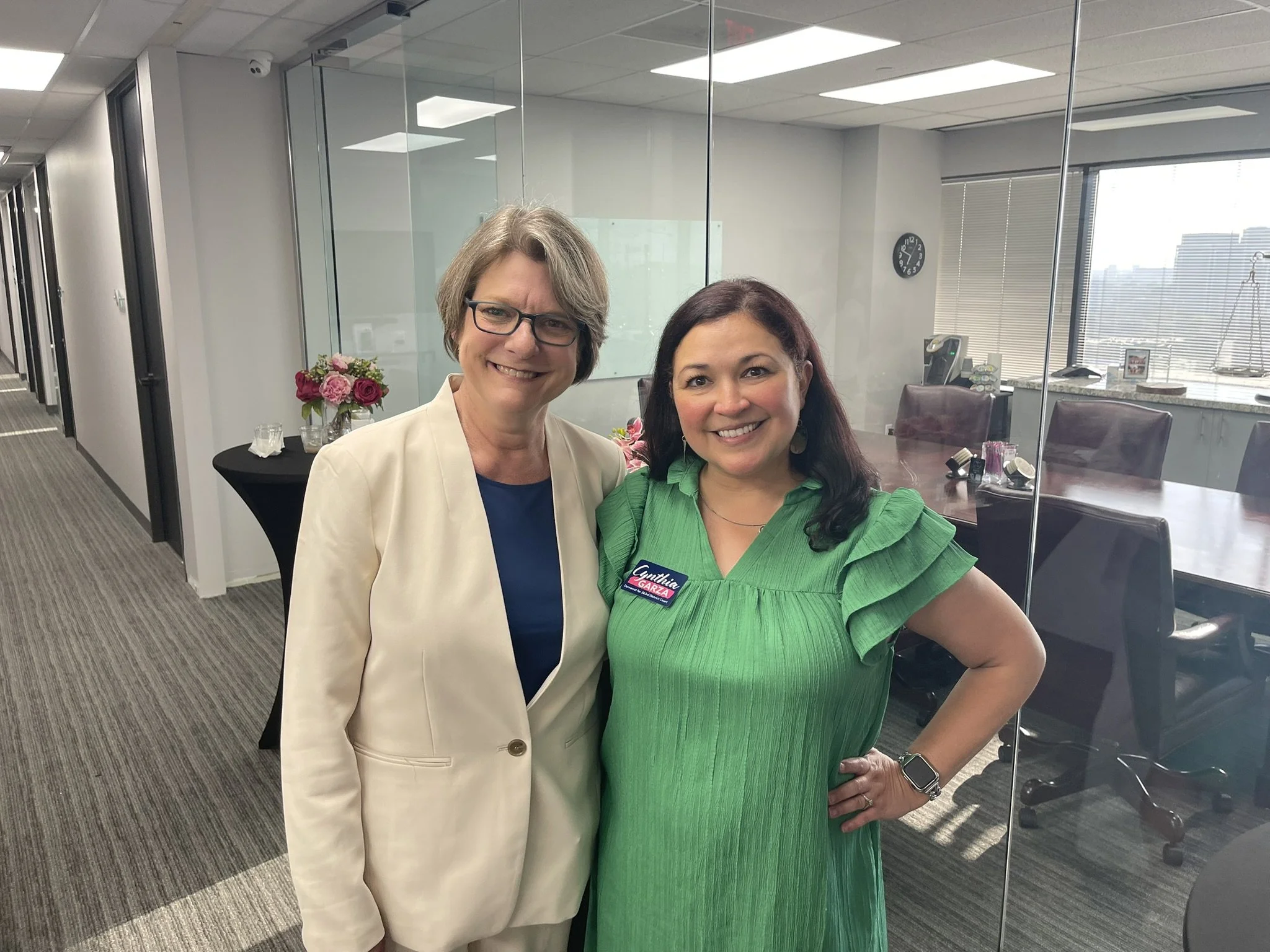 Two women are standing inside an office, smiling at the camera. One woman is wearing a beige blazer over a blue top, and the other is wearing a bright green dress with a name tag that reads 'Gretlin Garza.' There are flowers on a table behind them, a