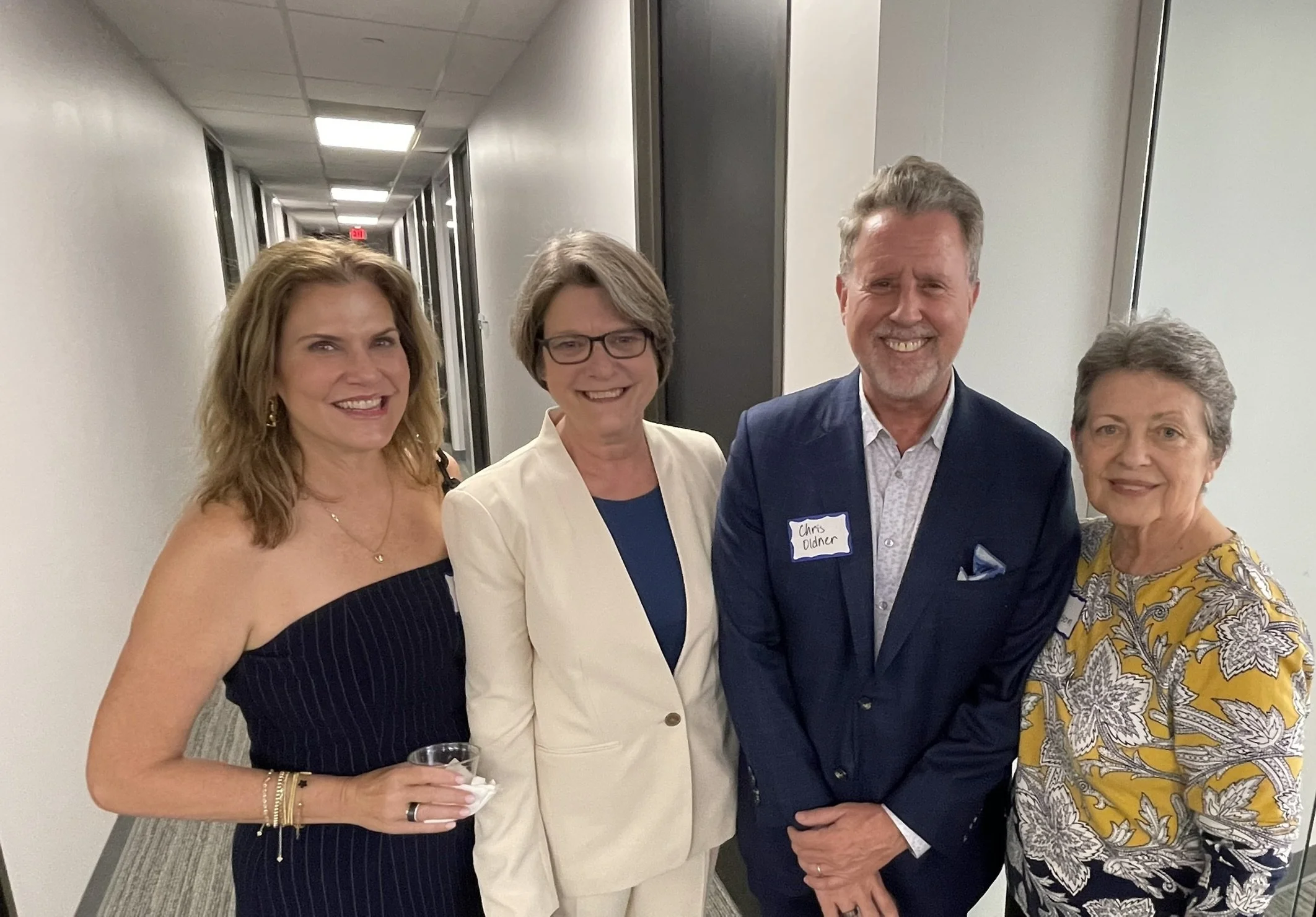 Four adults standing in a hallway, smiling for a photo. The woman on the far left is holding a drink, wearing a strapless black dress. The second woman from the left is wearing glasses and a white blazer. The man in the middle is wearing a blue suit 