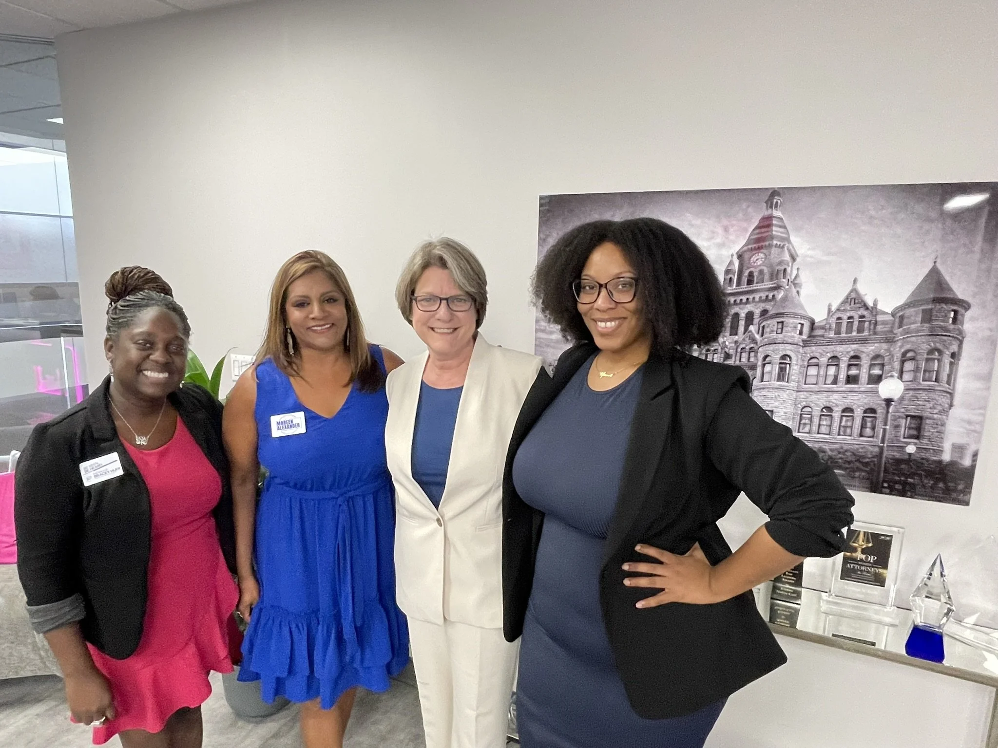 Four women standing together smiling in an indoor setting, with a large black-and-white photograph of a historic building on the wall behind them.