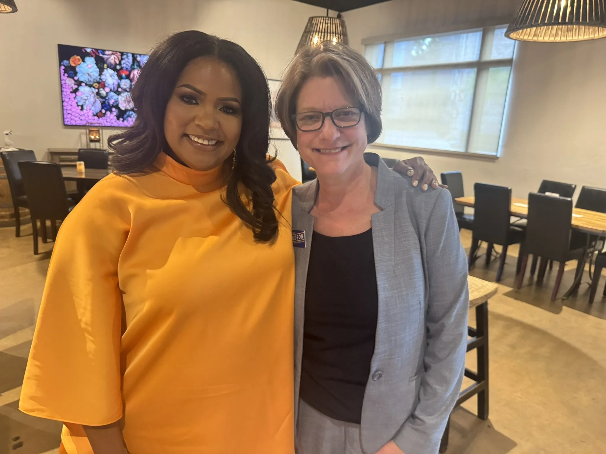 Two women standing together in a room, smiling for the camera. The woman on the left is wearing a bright yellow dress, and the woman on the right is wearing a gray blazer and glasses, with her arm around the other woman.