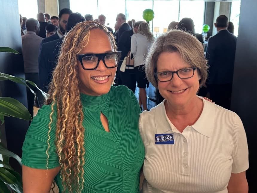 Two women smiling at an indoor social or networking event, with a crowd of people in business attire in the background.