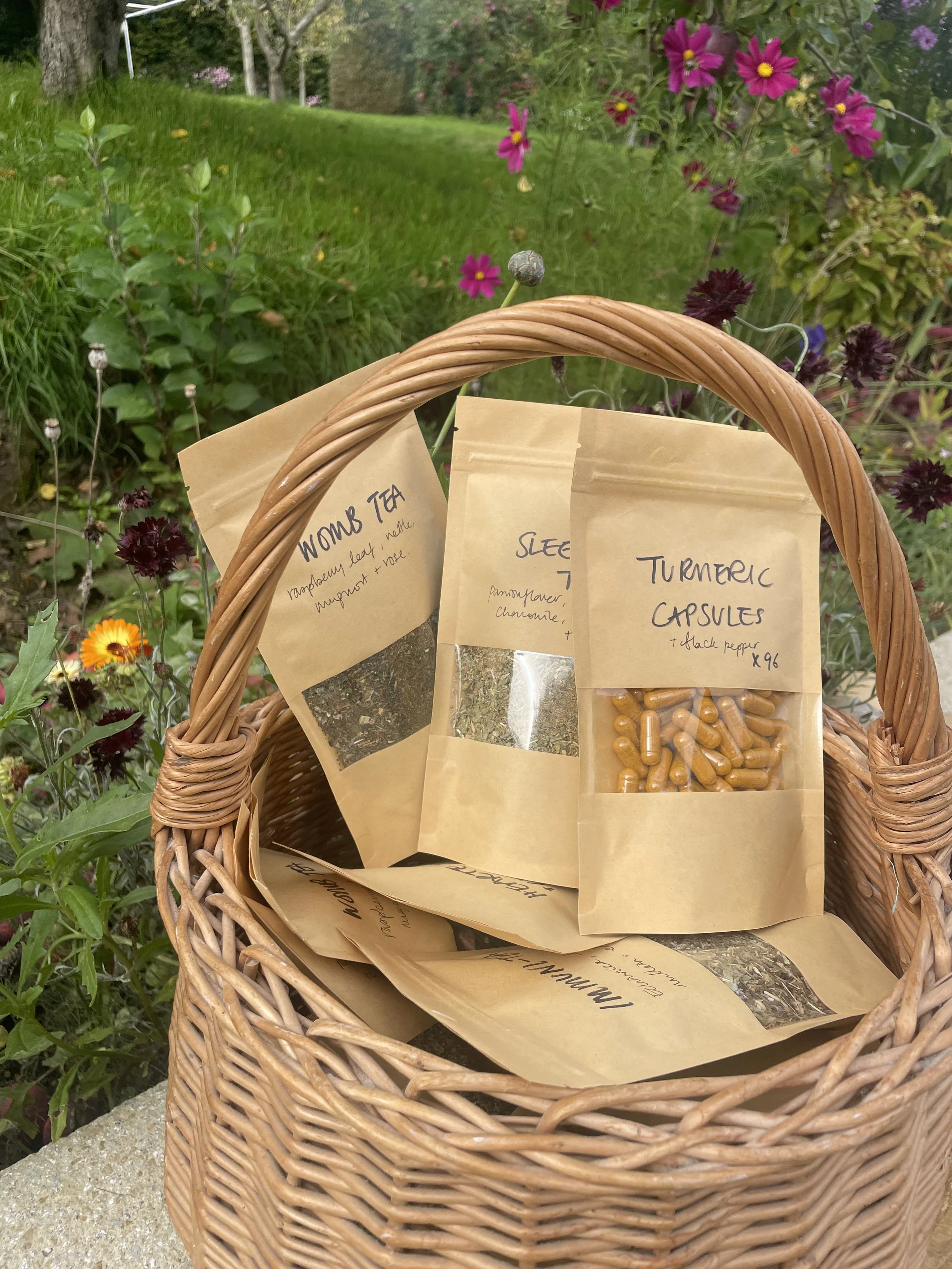 A wicker basket containing paper packets labeled 'Womb Tea,' 'Sleep,' and 'Turmeric Capsules,' with a garden of colorful flowers and greenery in the background.