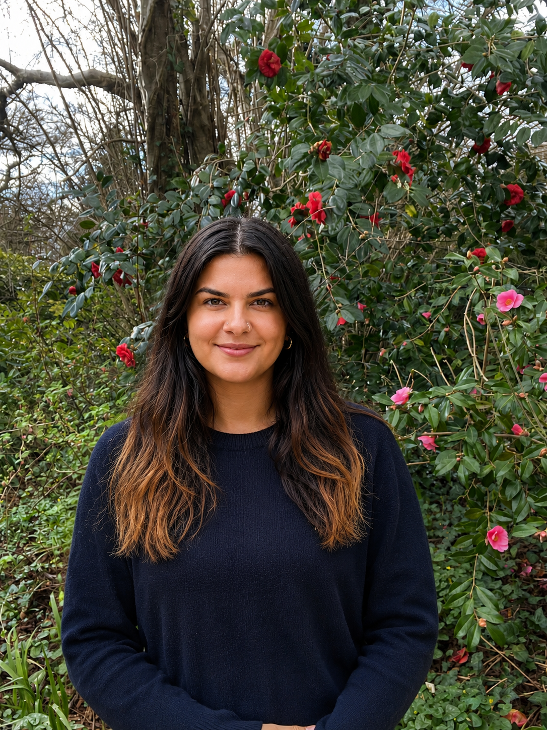 A young woman with dark brown hair with lighter tips, wearing a black sweater, standing in front of green bushes with red and pink flowers.