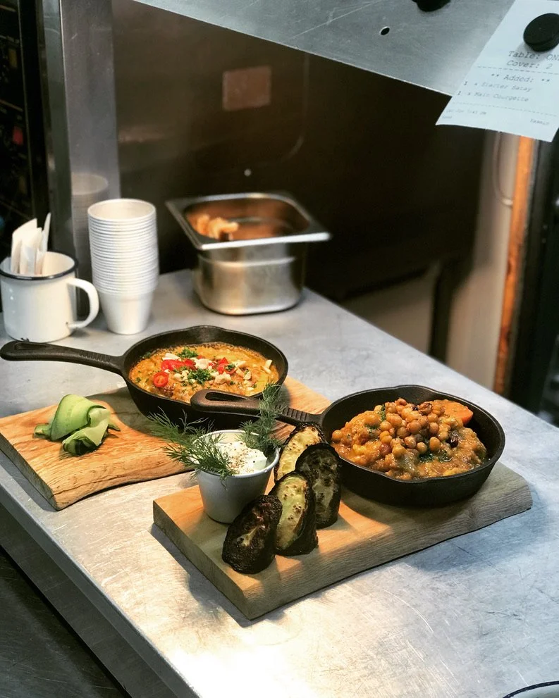 Two cast iron skillets with Indian curry dishes on a wooden serving board, accompanied by grilled eggplant slices, a small bowl of herbs, and cucumber slices, on a metal kitchen countertop.