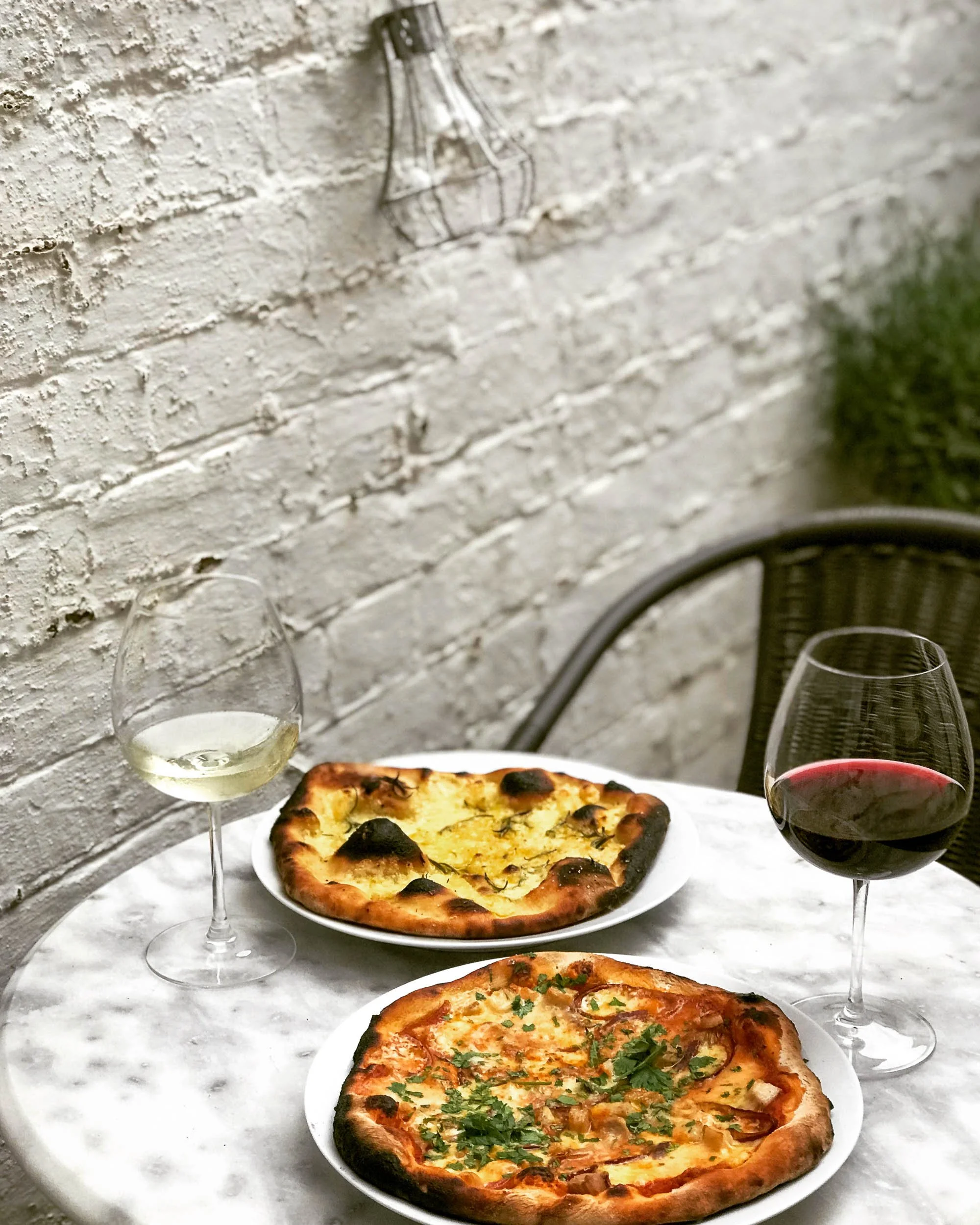 Two glasses of white and red wine on a table with two pizzas, one with cheese and herbs and the other with toppings, against a white brick wall background.