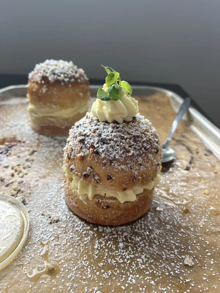Two crème brûlée profiteroles topped with whipped cream, a sprig of microgreens, and powdered sugar, served on a tray with caramel sauce and a small dish of cream.