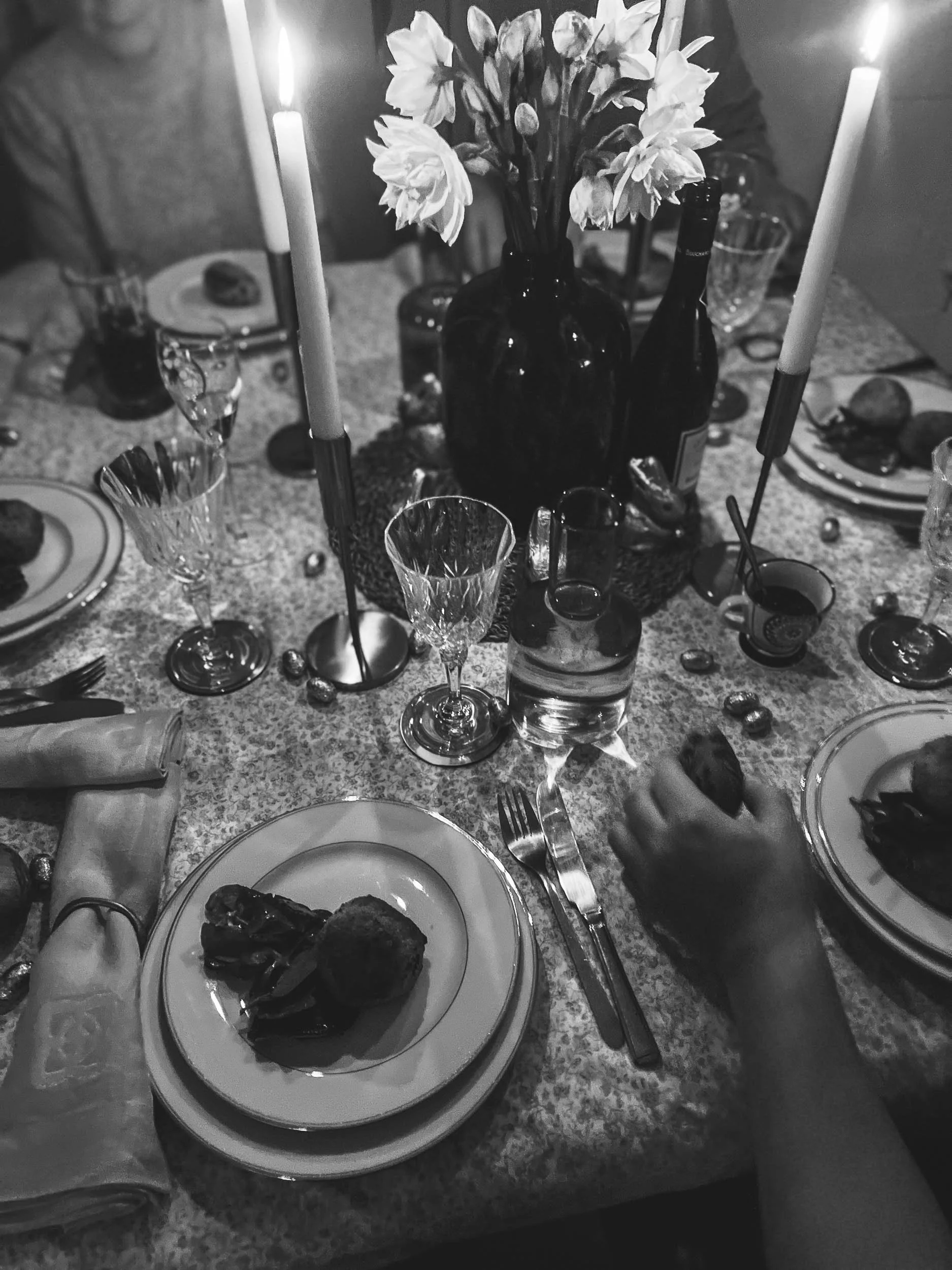 A black and white photo of a dinner table with plates, glasses, candles, and a centerpiece of flowers in a vase.