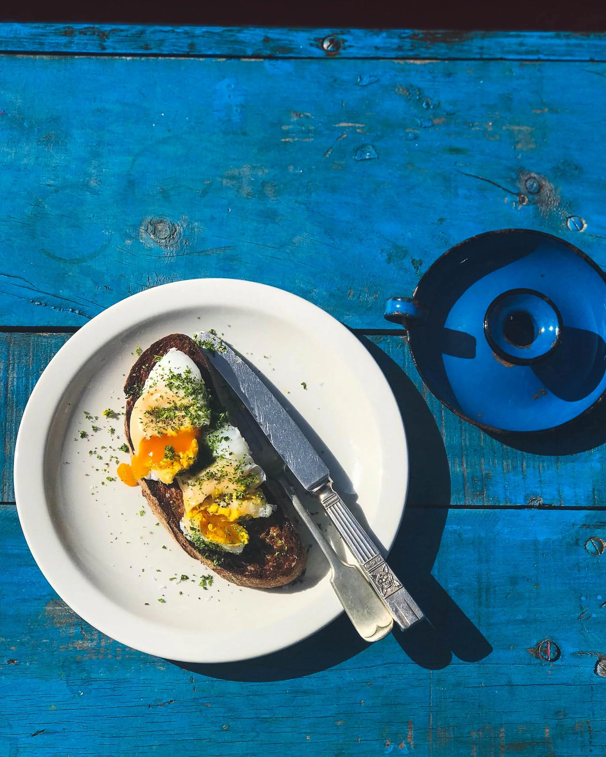 Poached egg and hollandaise sauce on toasted bread on a white plate with a butter knife, on a bright blue wooden table.