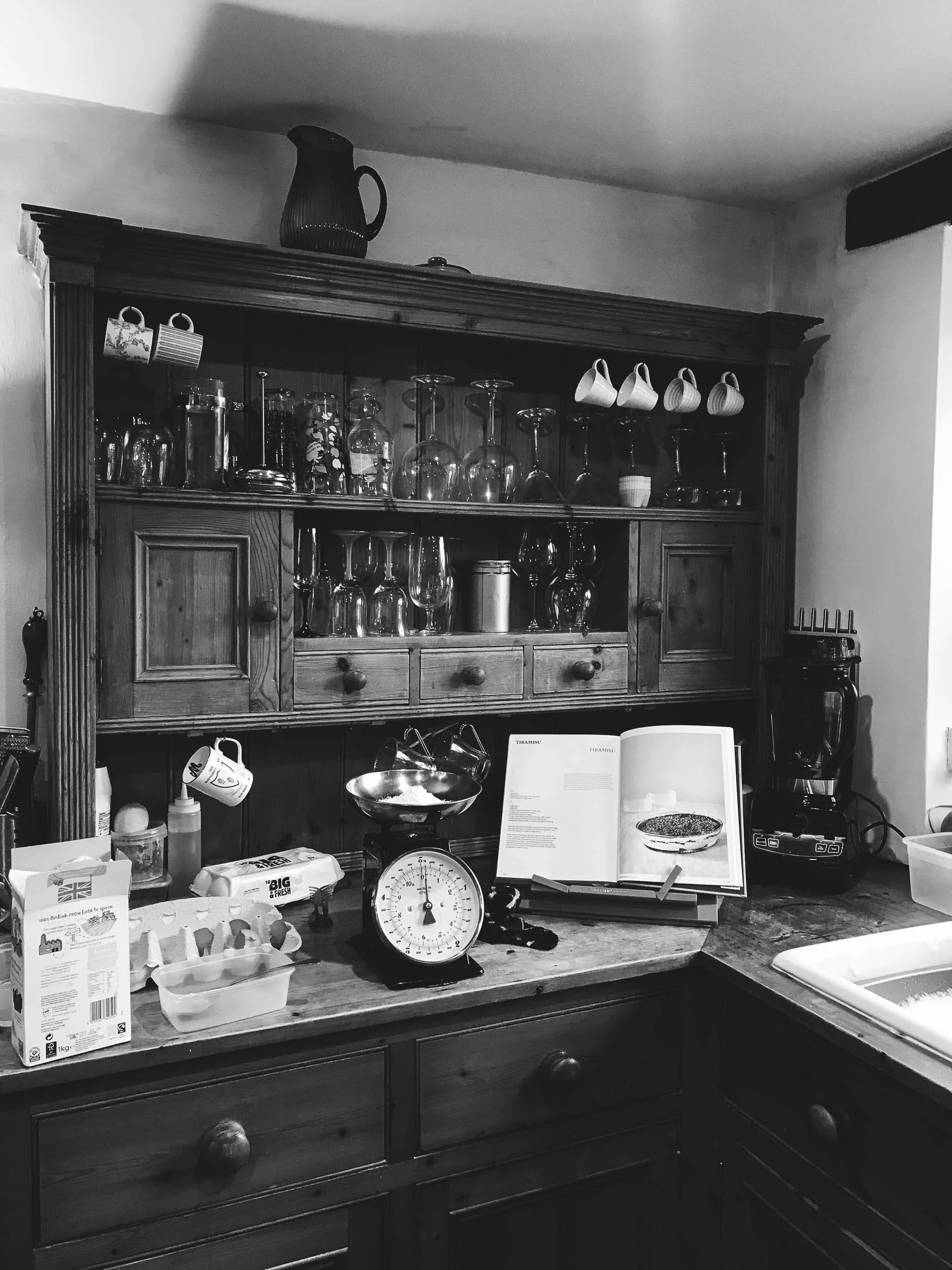 A kitchen counter with baking ingredients, a scale, and recipe book open, above a wooden cabinet with glassware and mugs.
