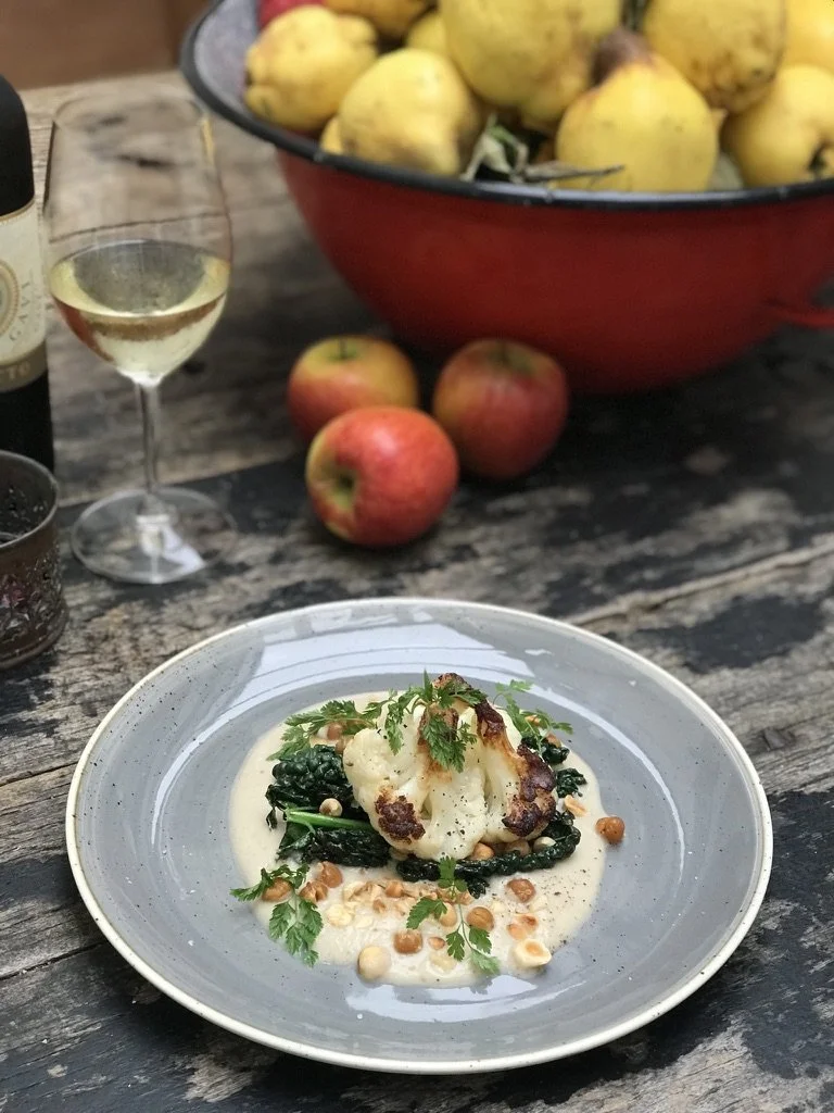 A gourmet dish featuring roasted cauliflower and sautéed greens, garnished with herbs and surrounded by a creamy sauce, on a rustic wooden table with apples, a glass of white wine, and a bowl of yellow summer squash in the background.